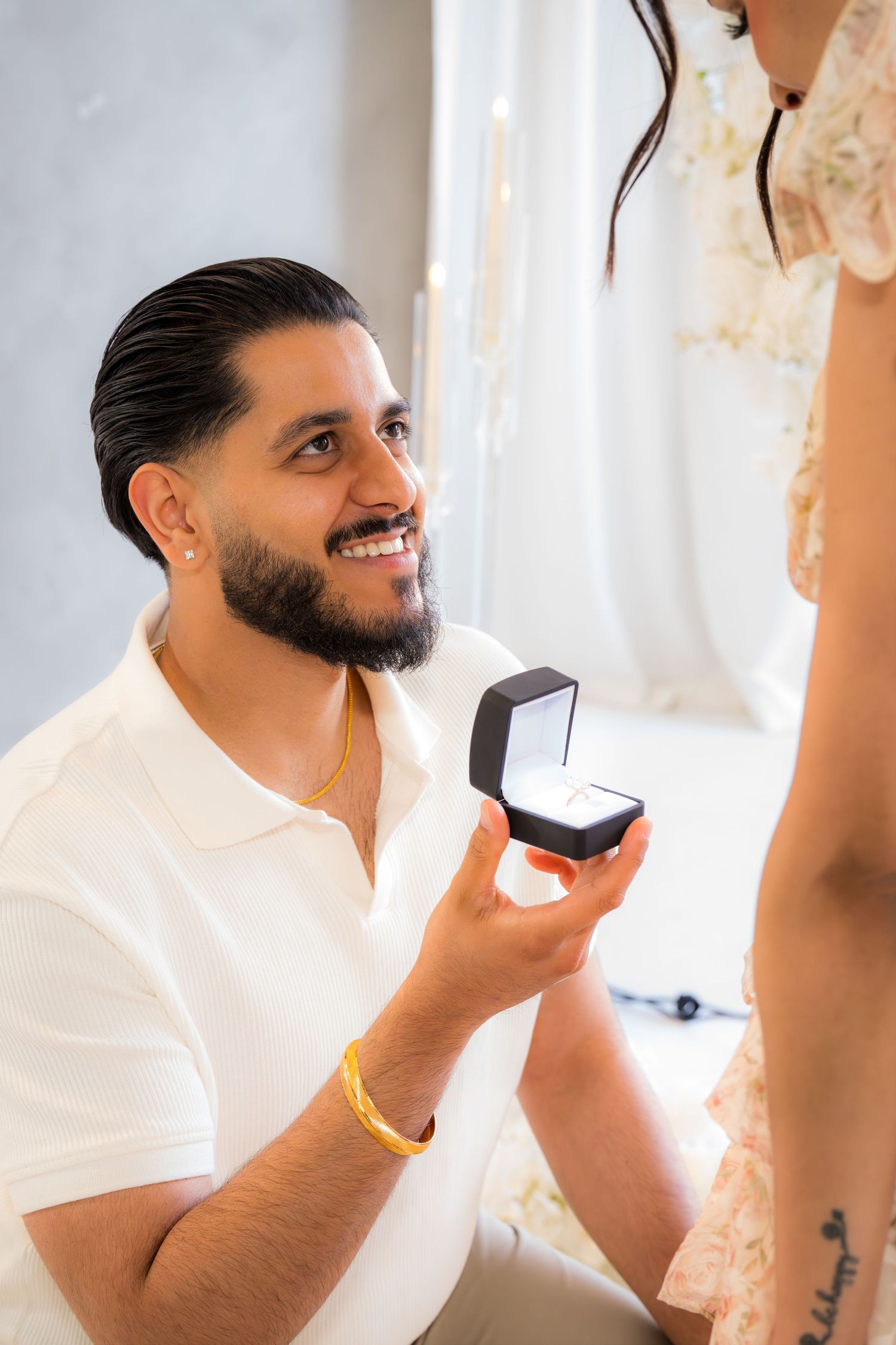 Man in white shirt kneeling on one knee presenting an engagement ring box while smiling up at his partner.