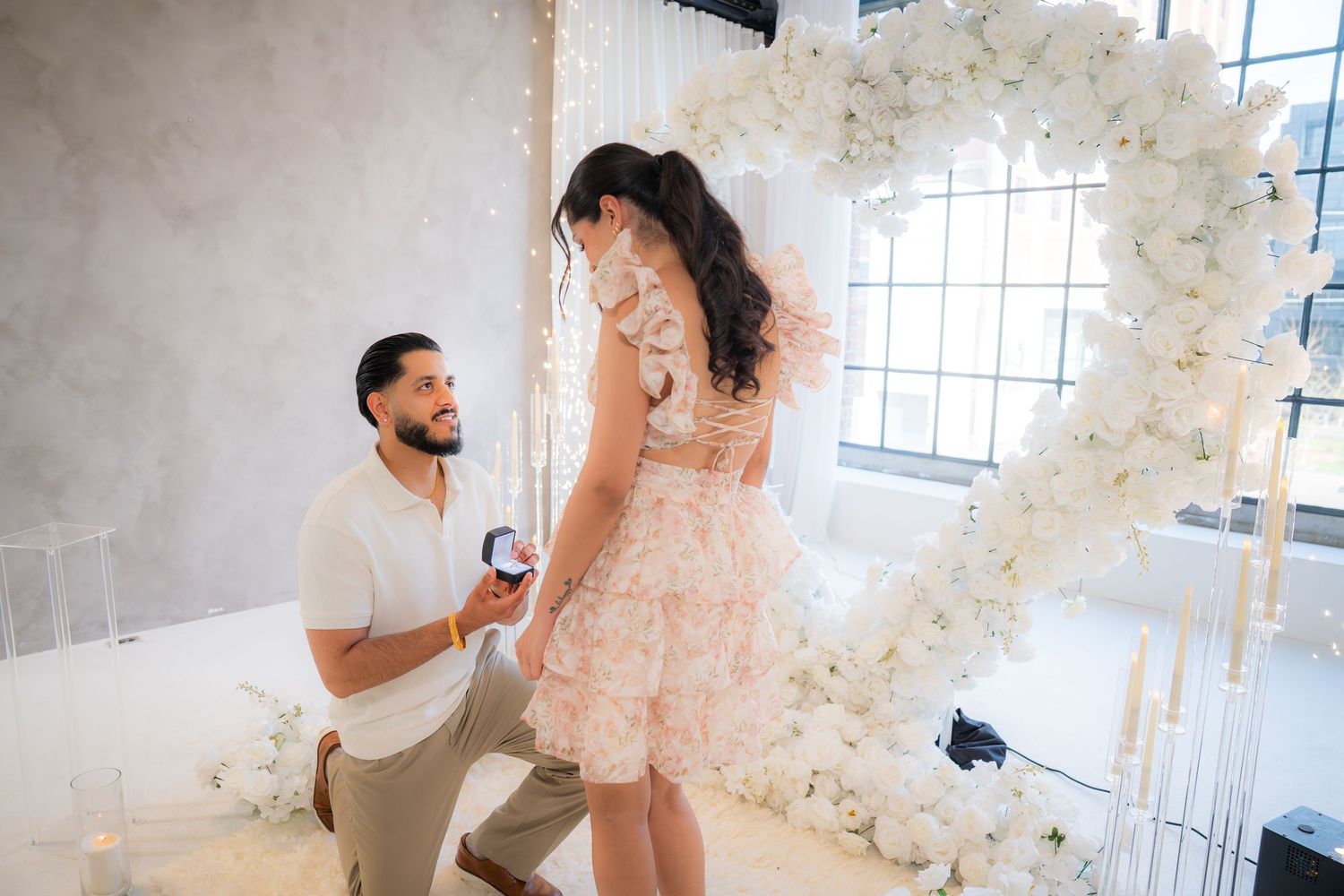 Man on one knee proposing to woman with a ring box in front of a white floral heart arch at an indoor venue.