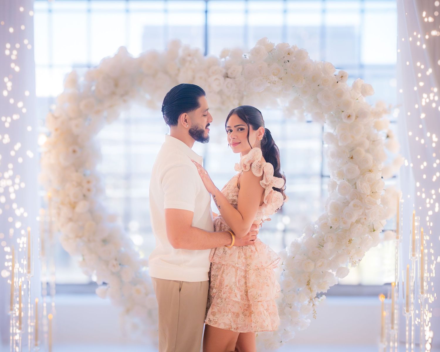 Couple holds each other tenderly inside a glowing white heart-shaped floral arch with fairy lights.