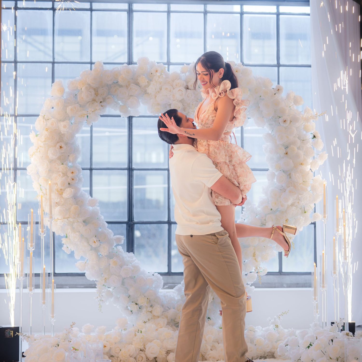 Joyful couple embracing in front of a stunning white floral heart arch with sparkler effects.