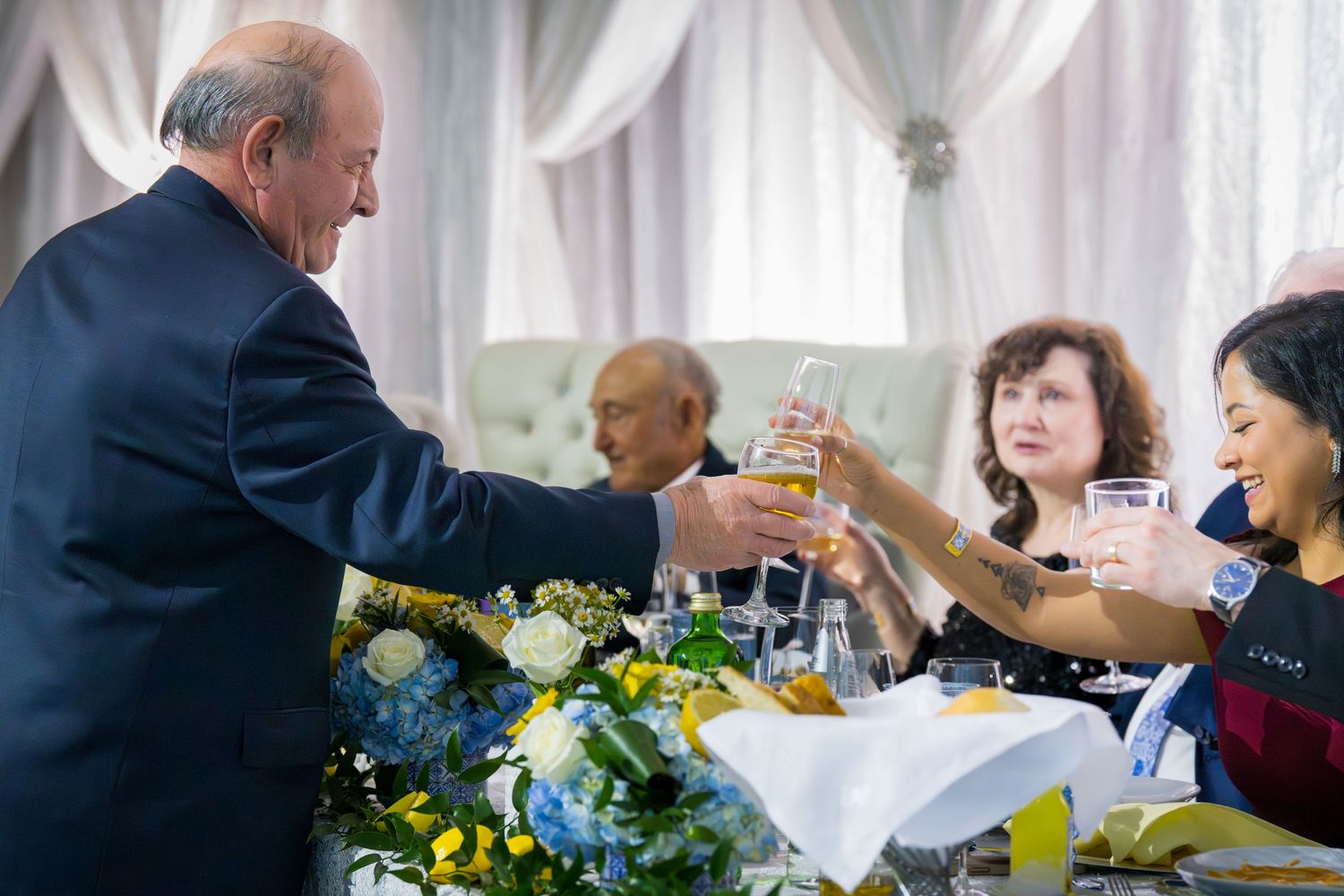 Guests raising glasses in a celebratory toast at an elegant banquet with blue and yellow floral centerpieces.