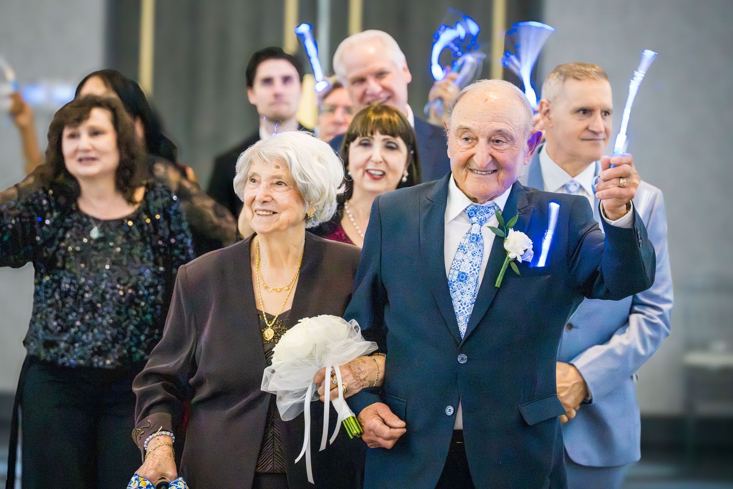Elderly couple walking arm in arm at a wedding celebration with guests cheering.