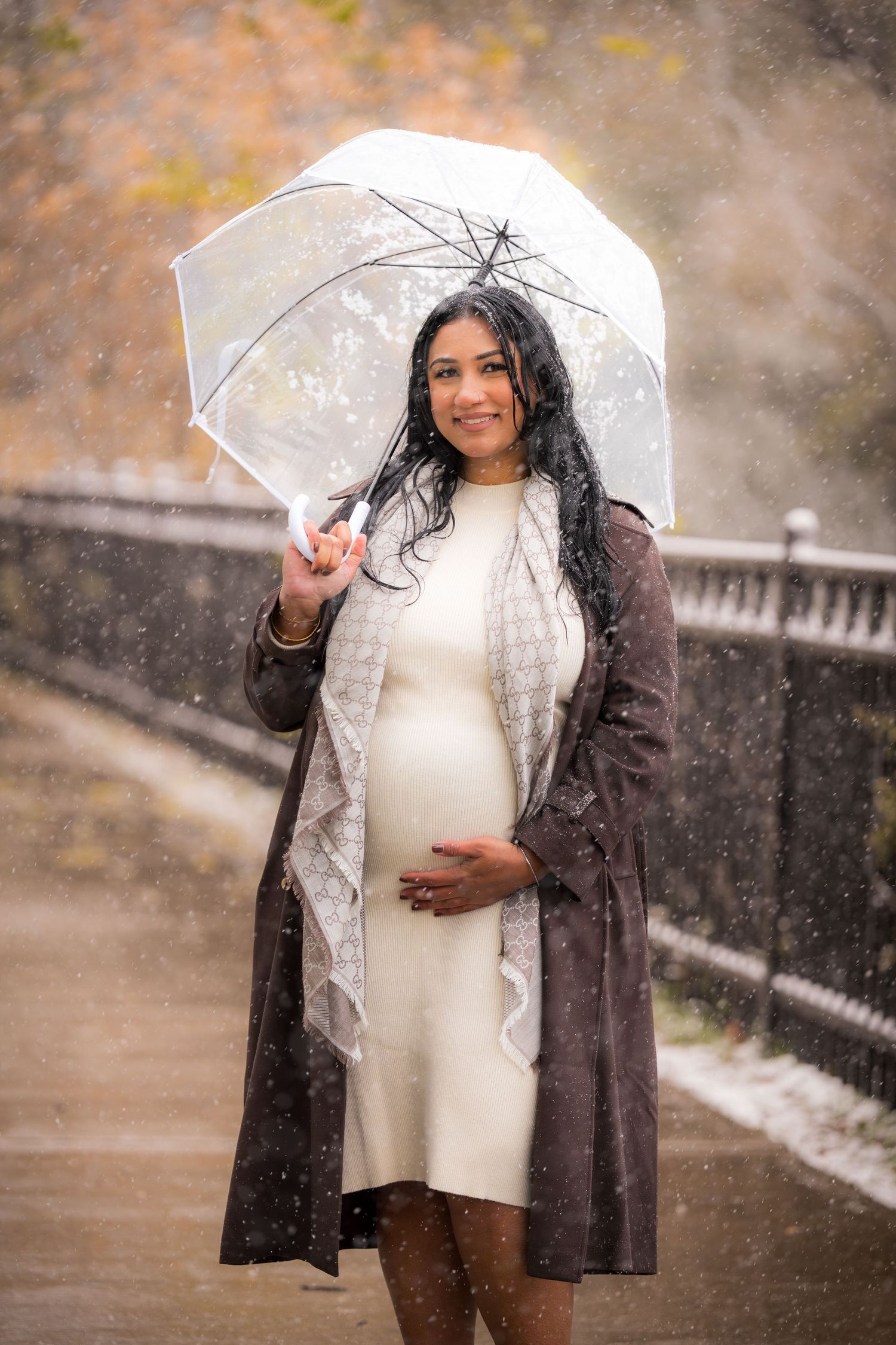 A person in a white sweater and dark coat poses with an umbrella during a snowy photoshoot.