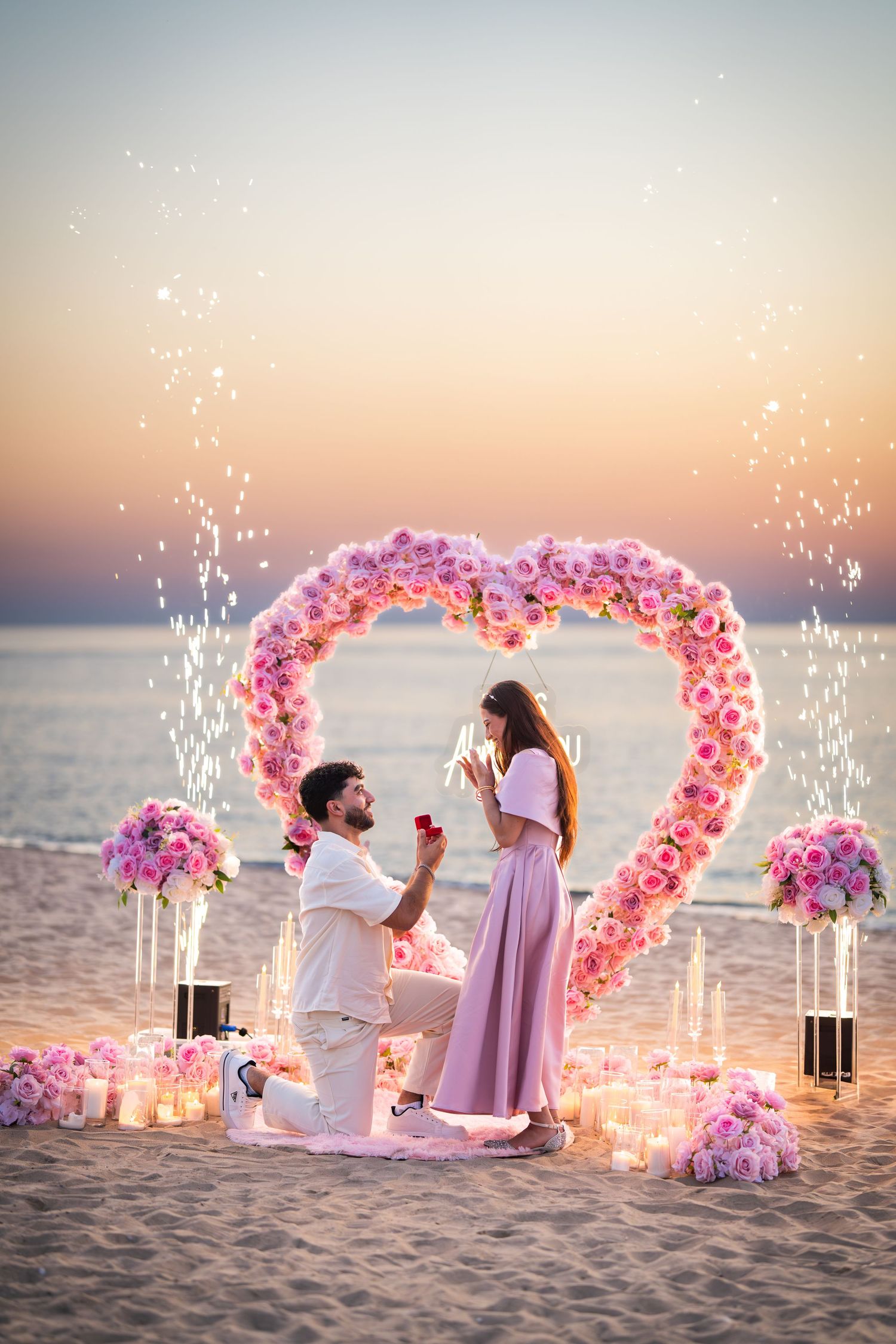 Beach proposal moment captured under a pink floral arch with sparkling fireworks at sunset.