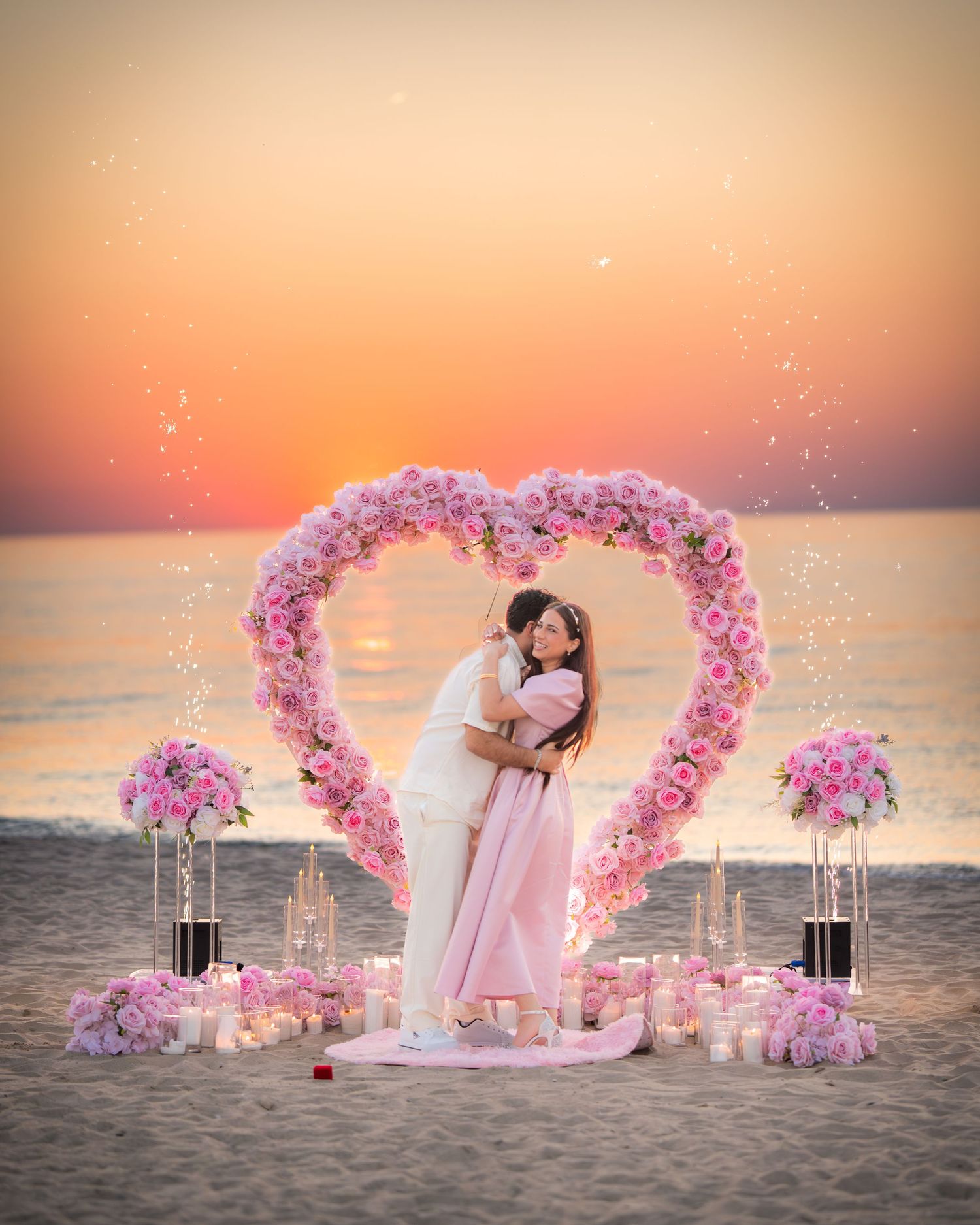 A sunset beach proposal with sparkling lights and a pink floral heart arch by the ocean.