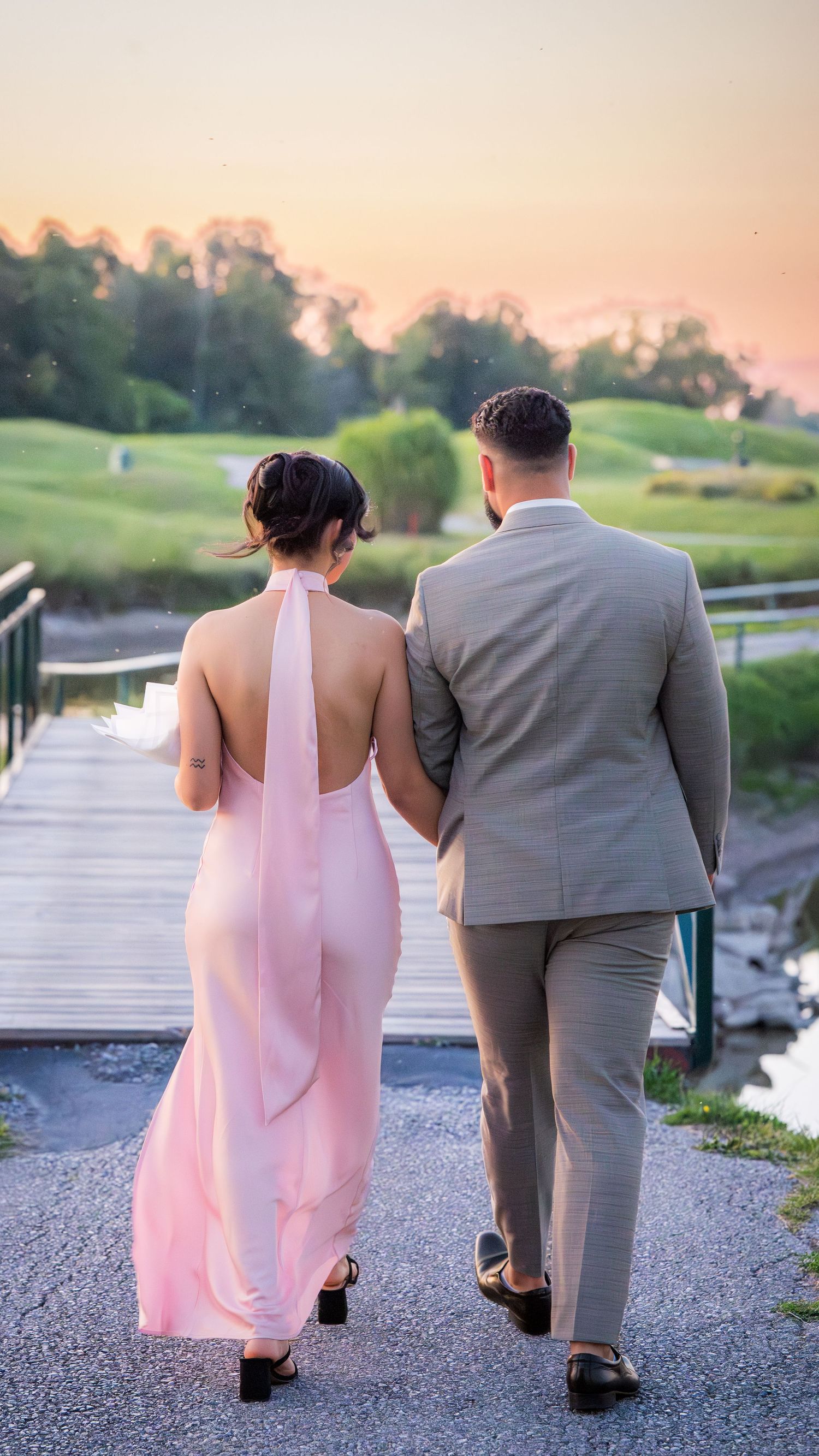 A couple walks away from camera on a path during sunset in formal attire.