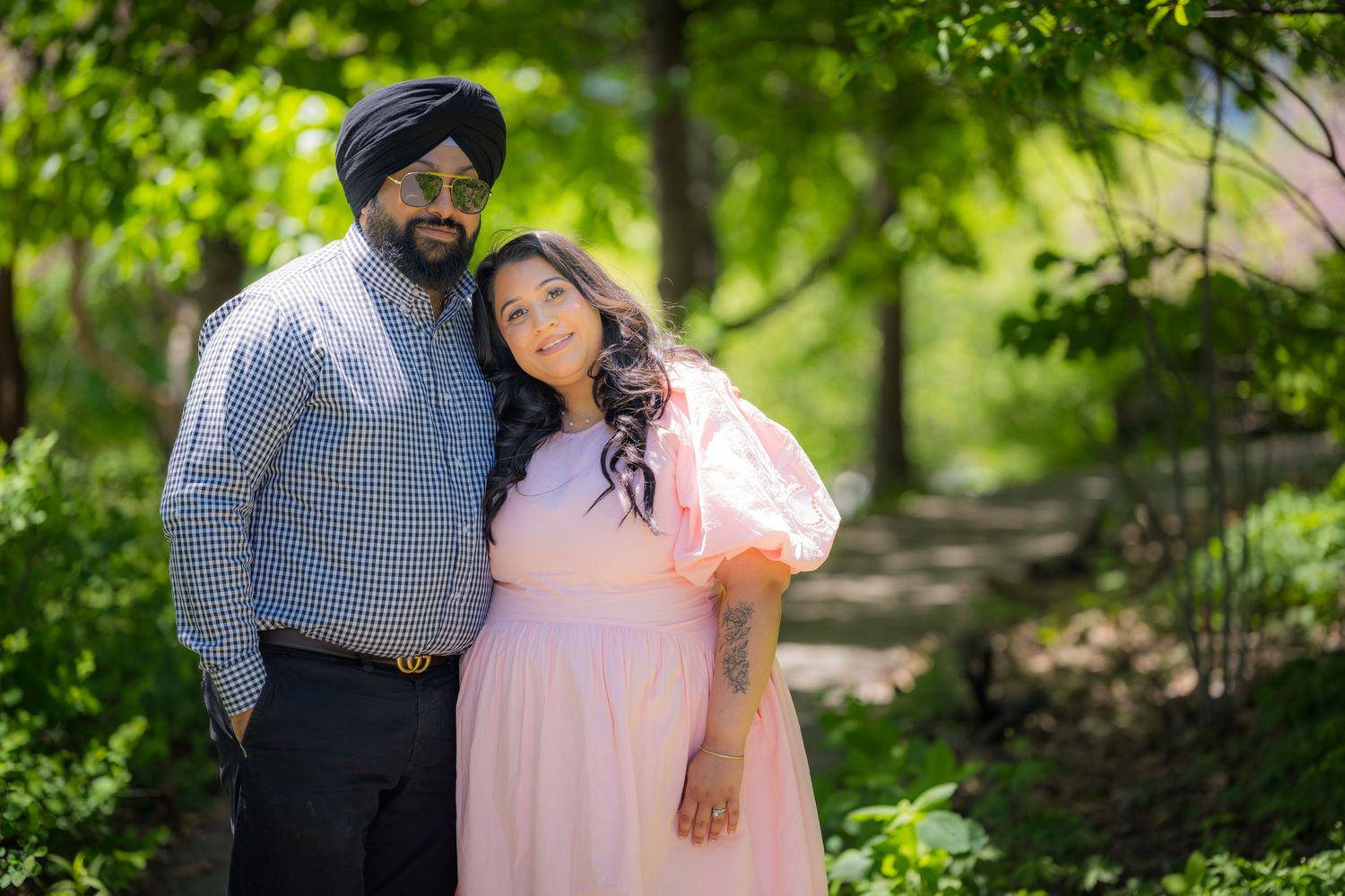 A Sikh couple poses outdoors in a lush green park, man in blue shirt and turban, woman in pink dress.