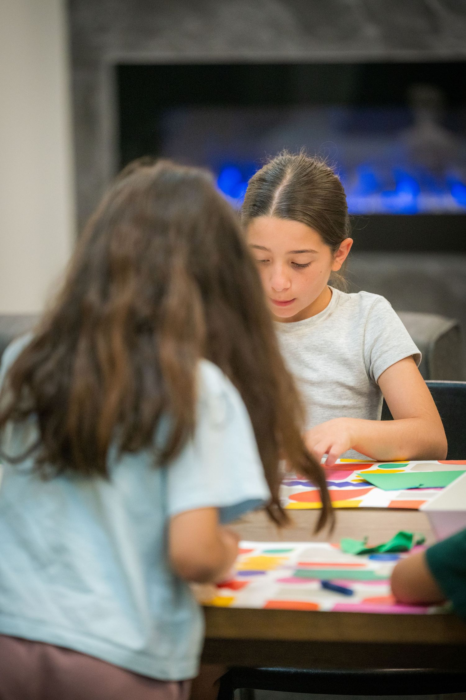 Two girls engage in colorful paper craft activity at a table near a fireplace.