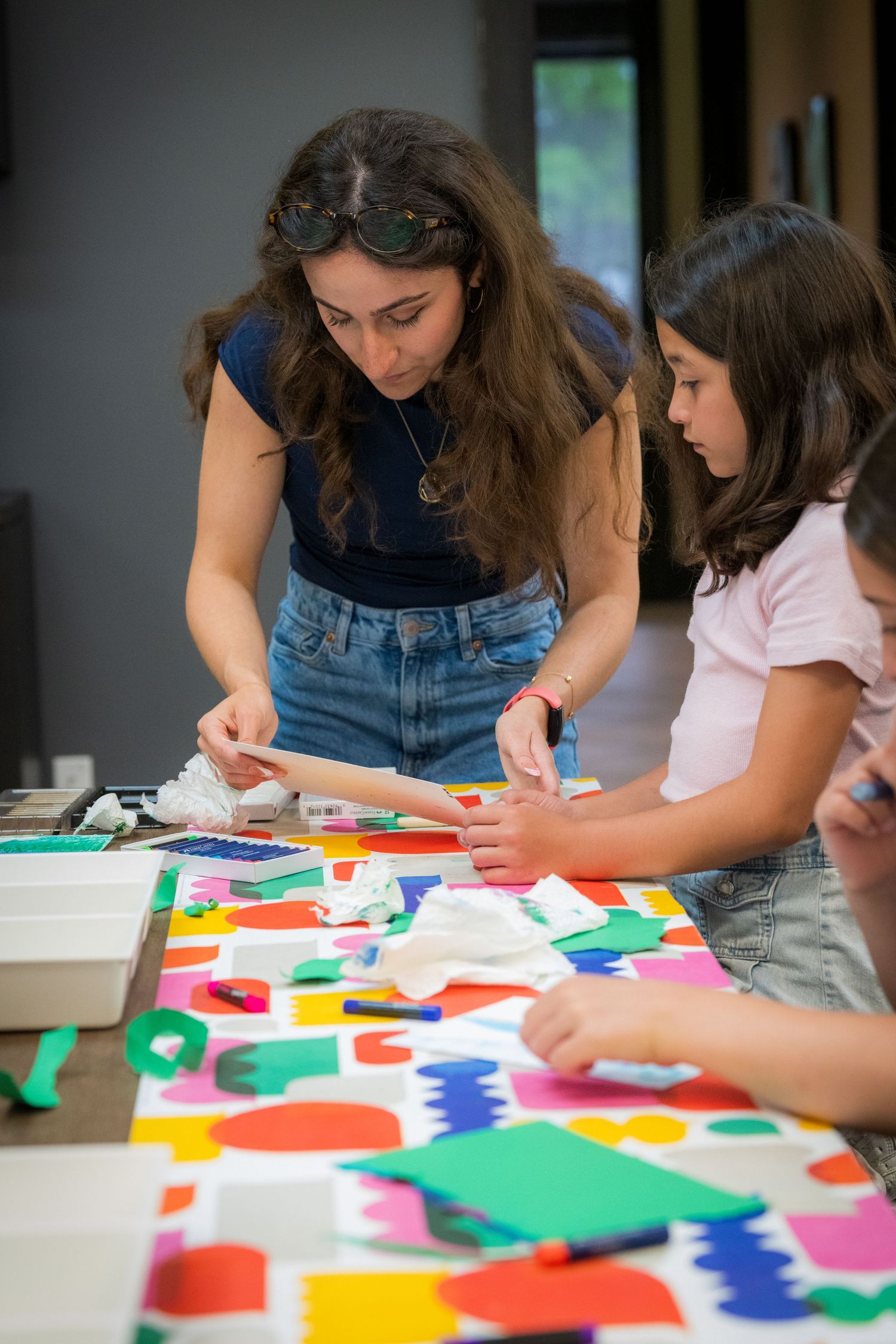 Art teacher and girl collaborate on vibrant colorful paper shape collage craft activity at table.