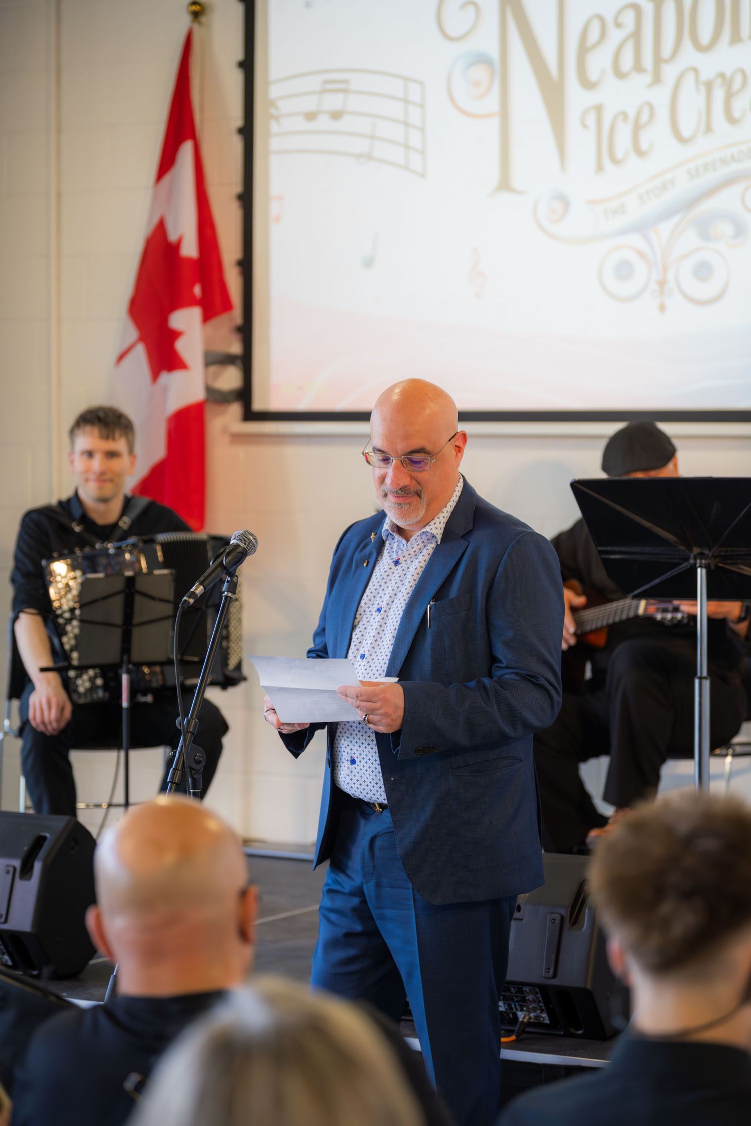 Bald man in blue blazer reads from paper at Neapolitan Ice Cream musical event with musicians performing behind him.