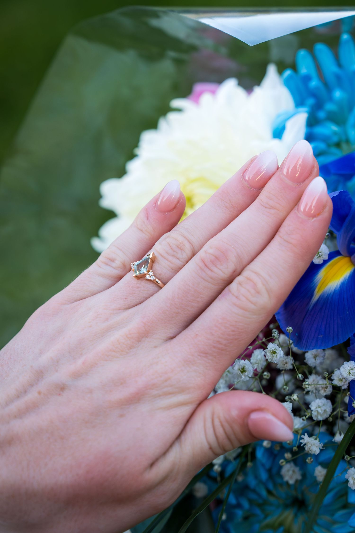 Close-up of a womans hand displaying a delicate gold engagement ring over a colorful bouquet.