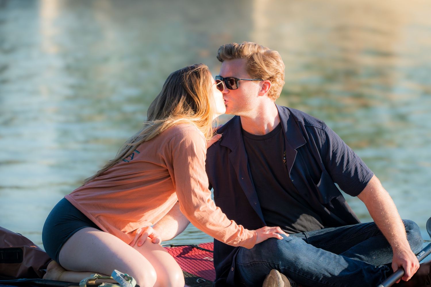 Young couple sharing a kiss while sitting on a dock by a sunlit lake.