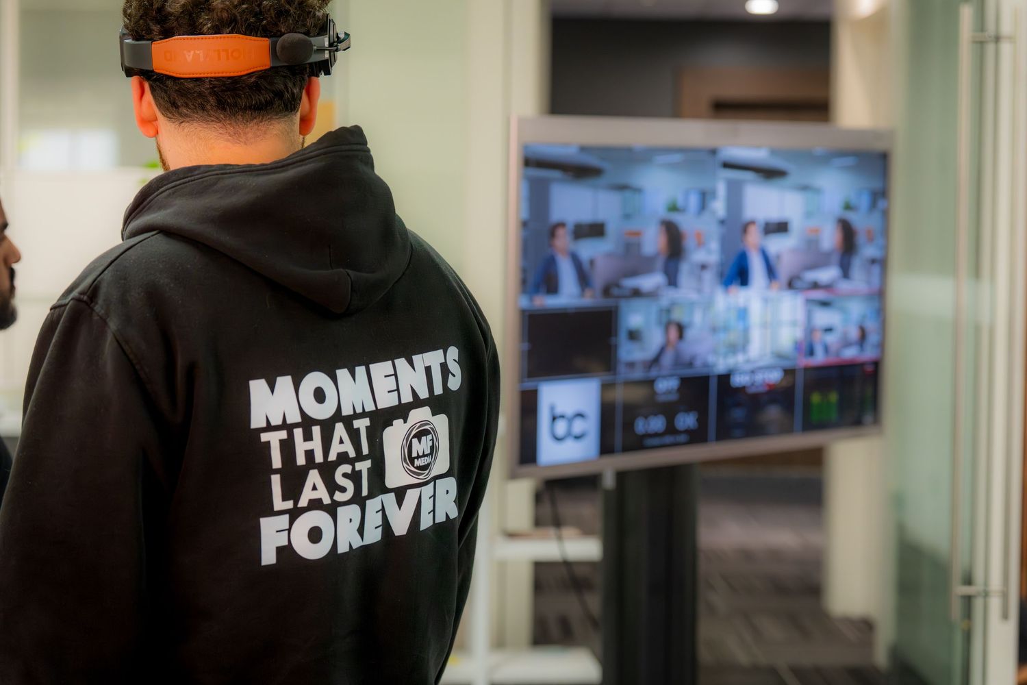 Person wearing black hoodie with 'Moments That Last Forever' text views a large monitor display at a tech event.