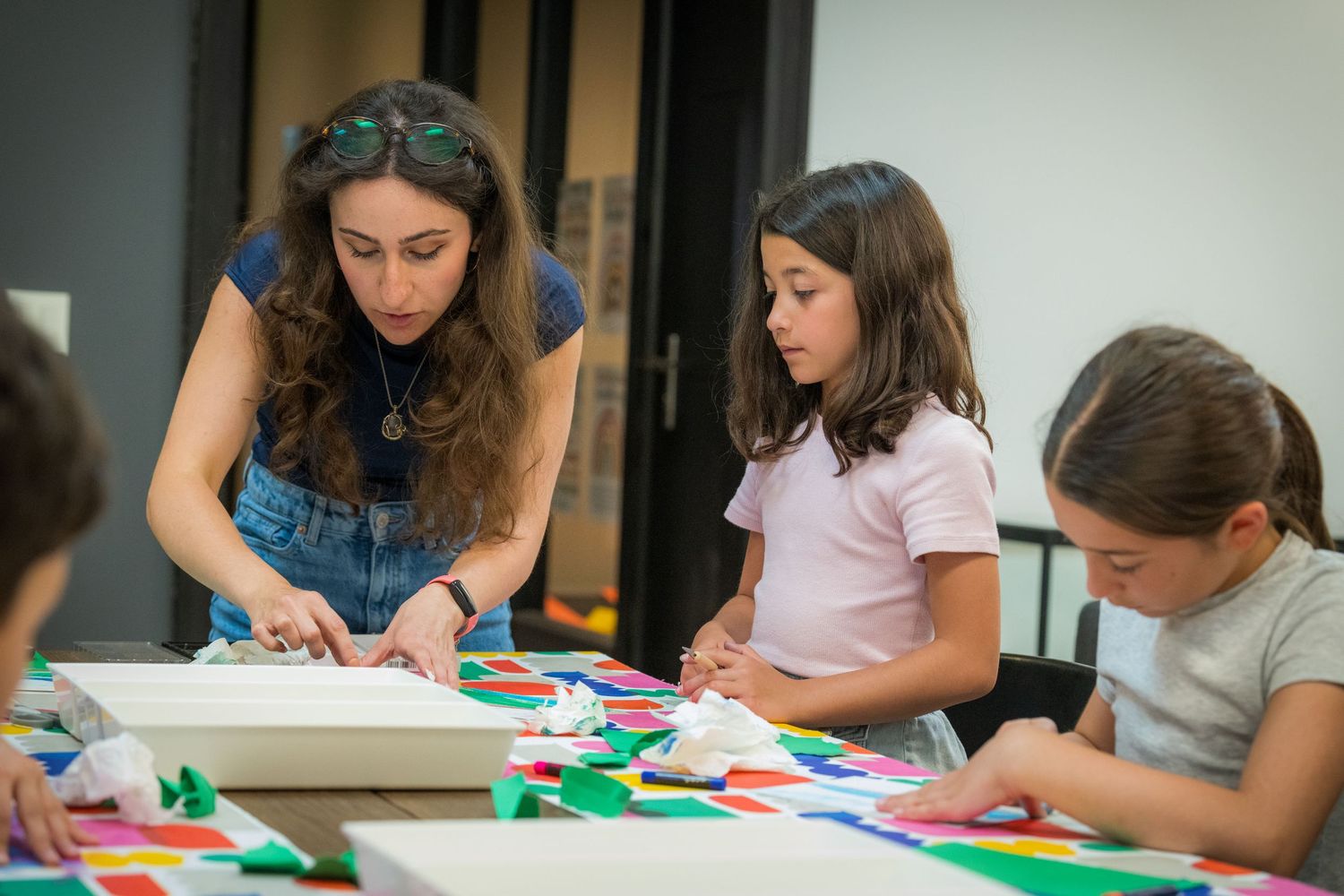A curly-haired instructor assists two young girls with a colorful paper art project indoors.