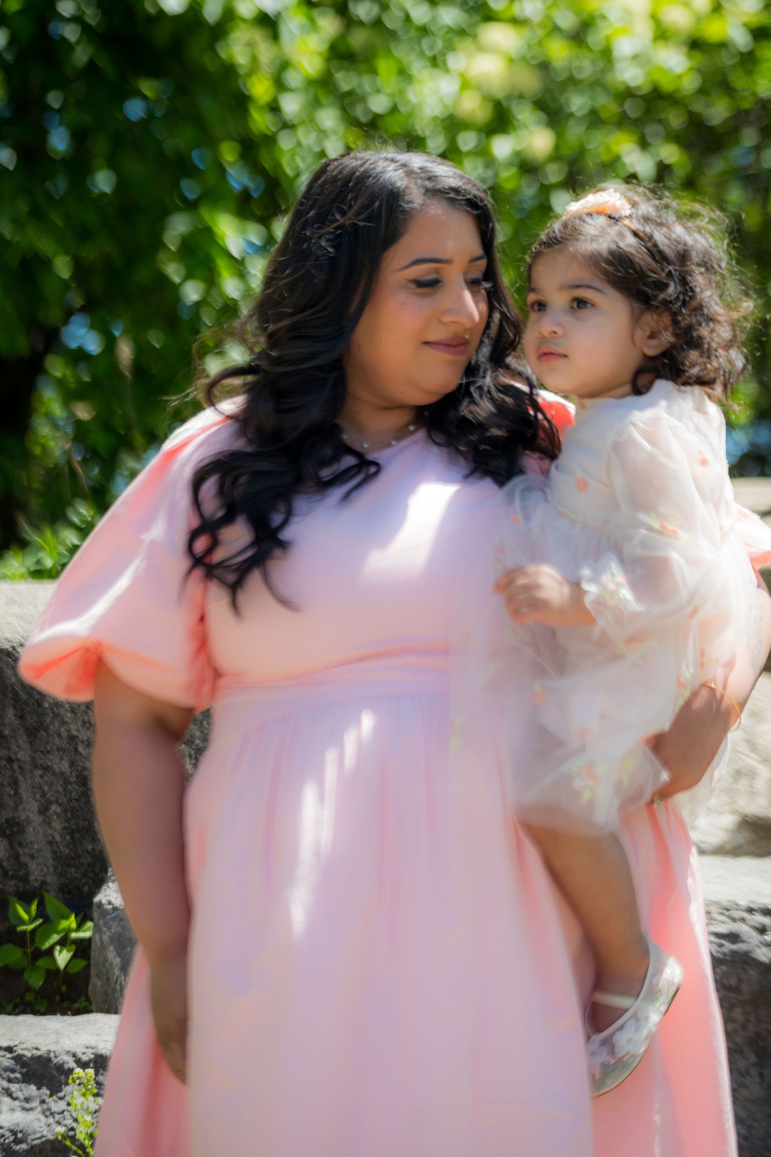Pregnant woman in pink dress holds toddler girl in white tulle dress in sunny park.