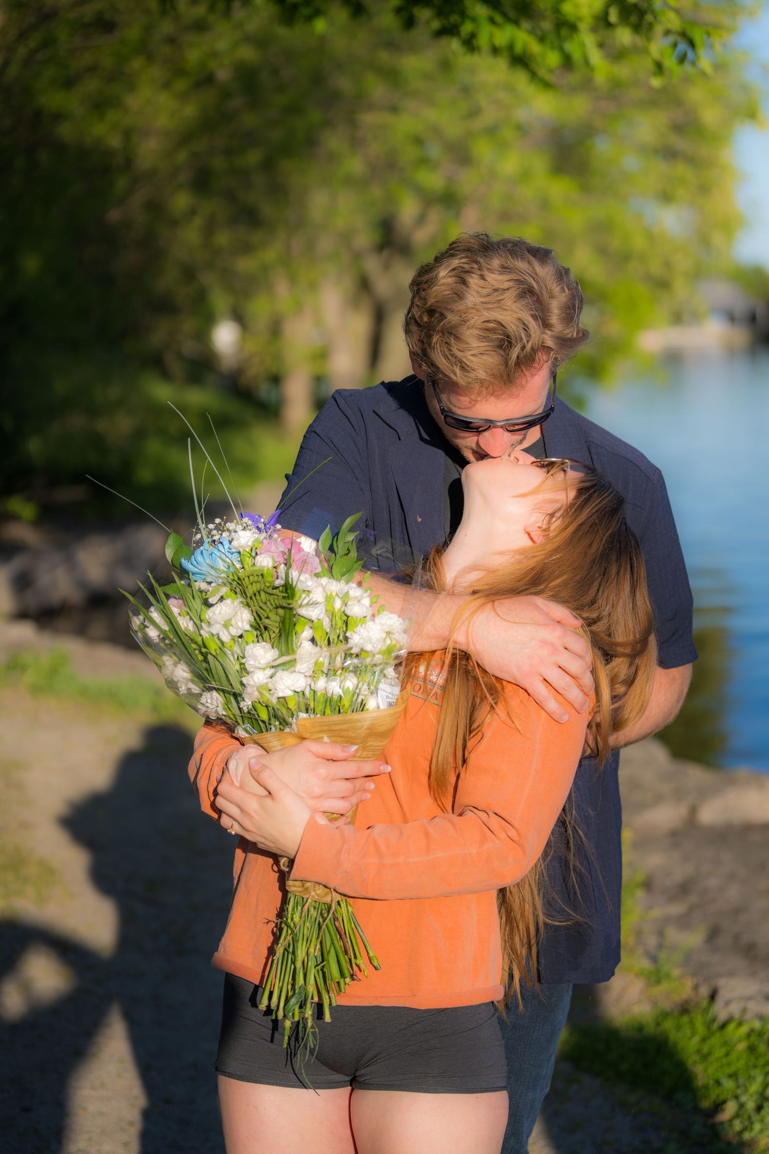 Couple sharing a kiss outdoors near a lake, woman holding a large bouquet of white flowers.