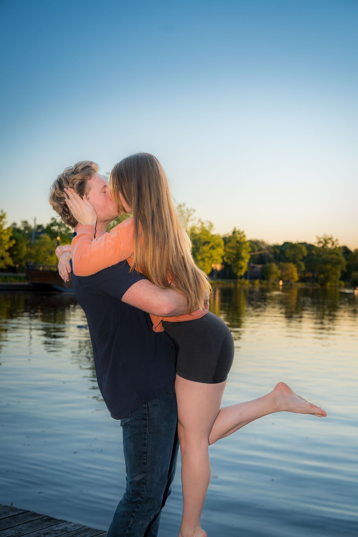 Couple sharing a romantic kiss by a lake at sunset, man lifting woman on a wooden dock.