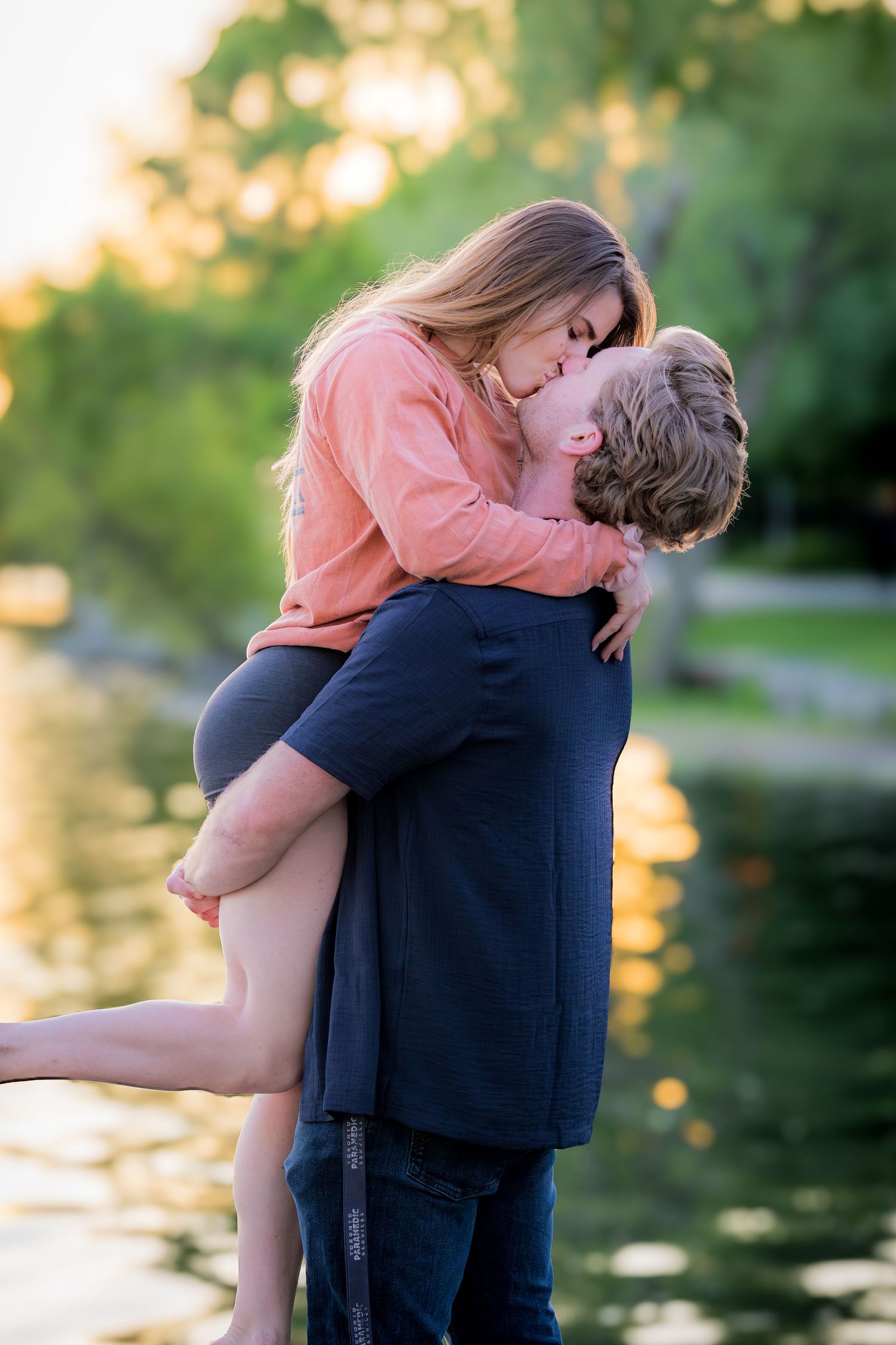 Man lifting and kissing woman near a sunlit lake surrounded by lush green trees at golden hour.