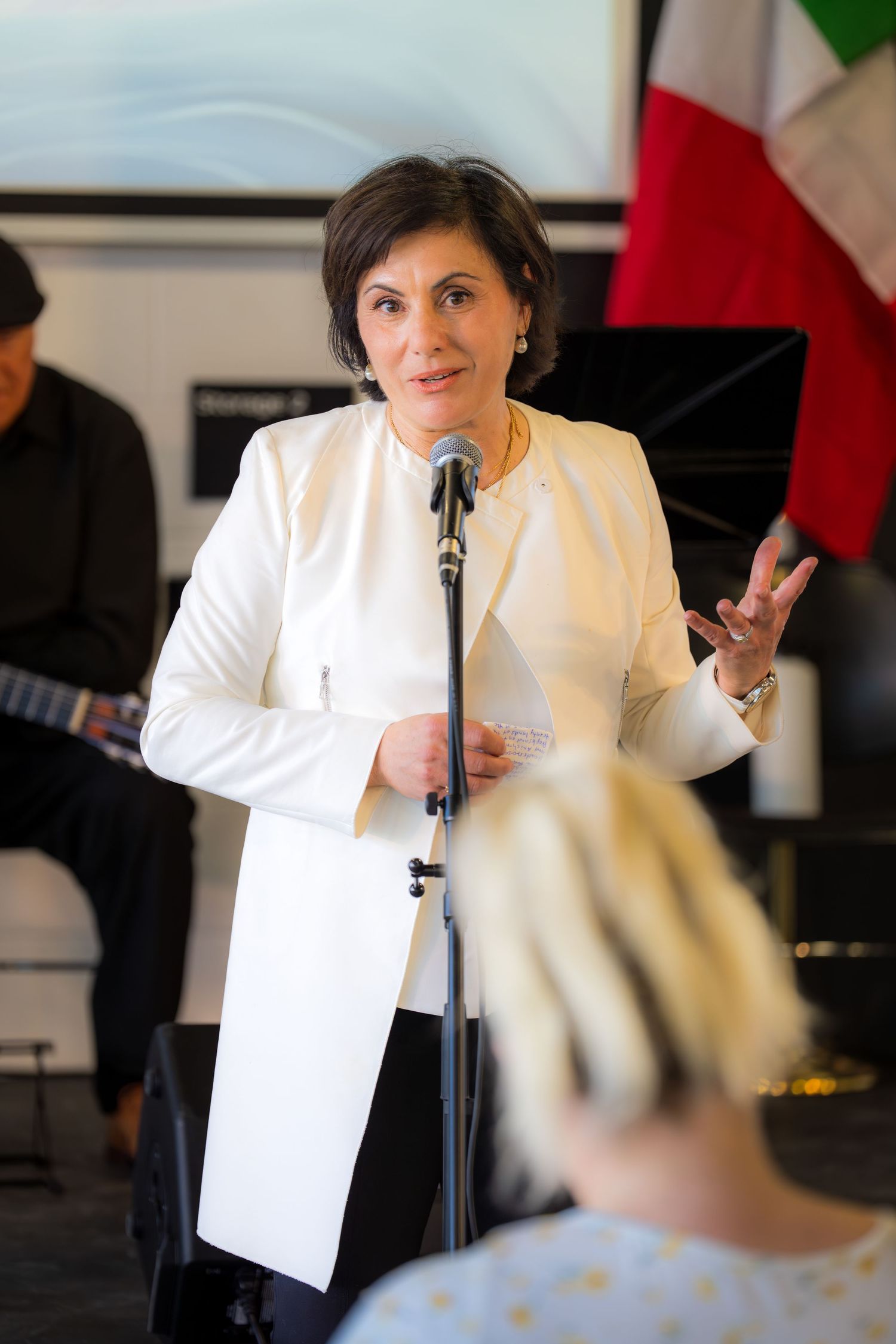 Elegant woman in white blazer addressing audience at microphone with Italian flag behind her.