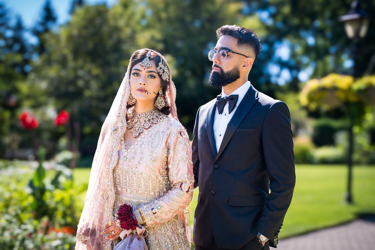 Newlywed couple poses together outdoors in a garden setting with lush greenery and trees in the background.