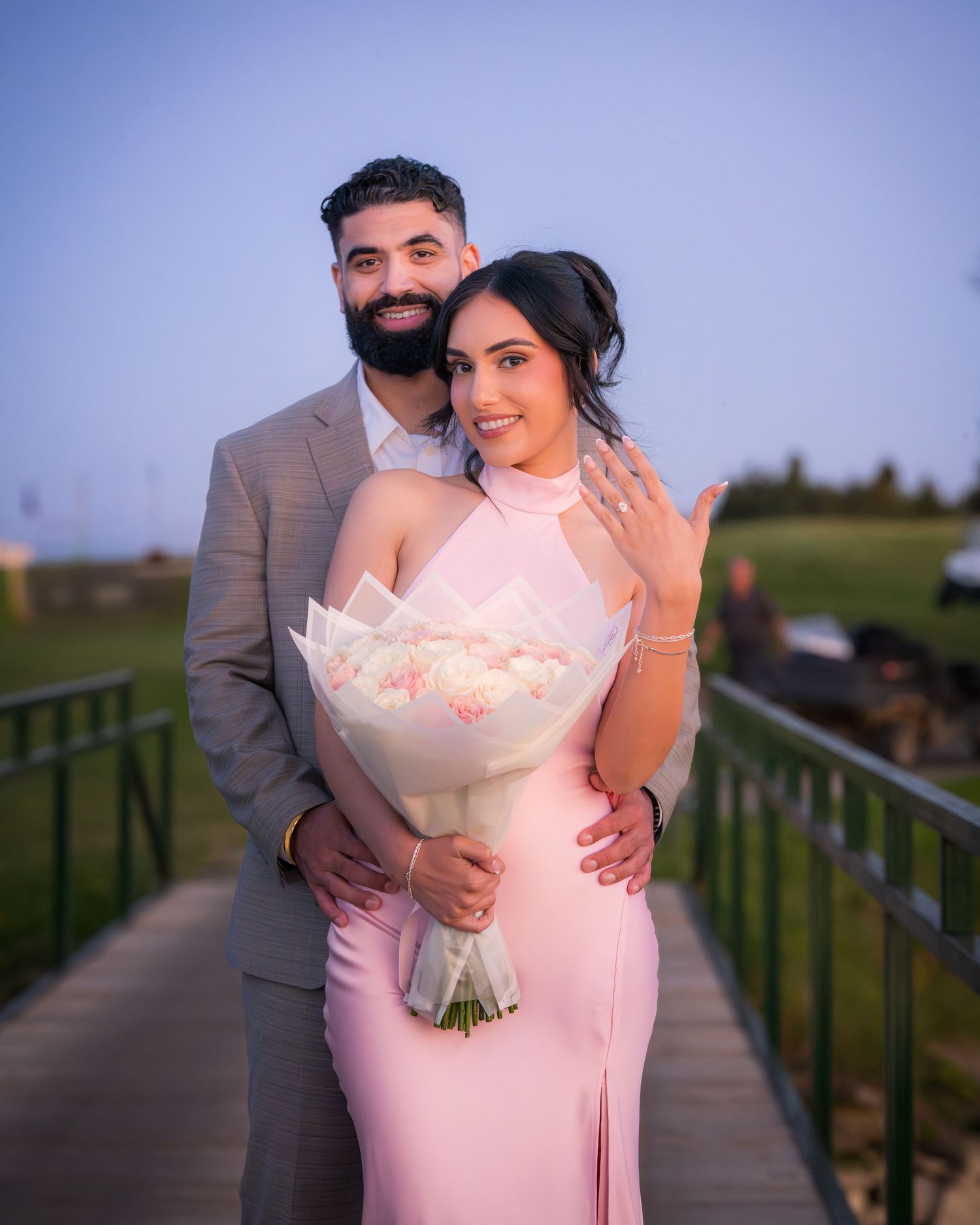 A romantic sunset photoshoot of a couple embracing, with one wearing a pink dress and holding flowers.