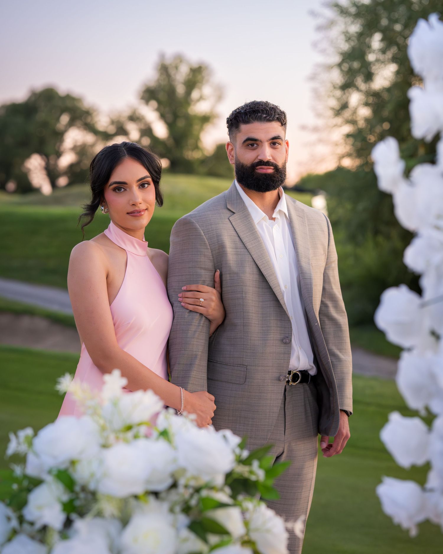 Romantic couple poses by white floral arch during sunset on golf course.