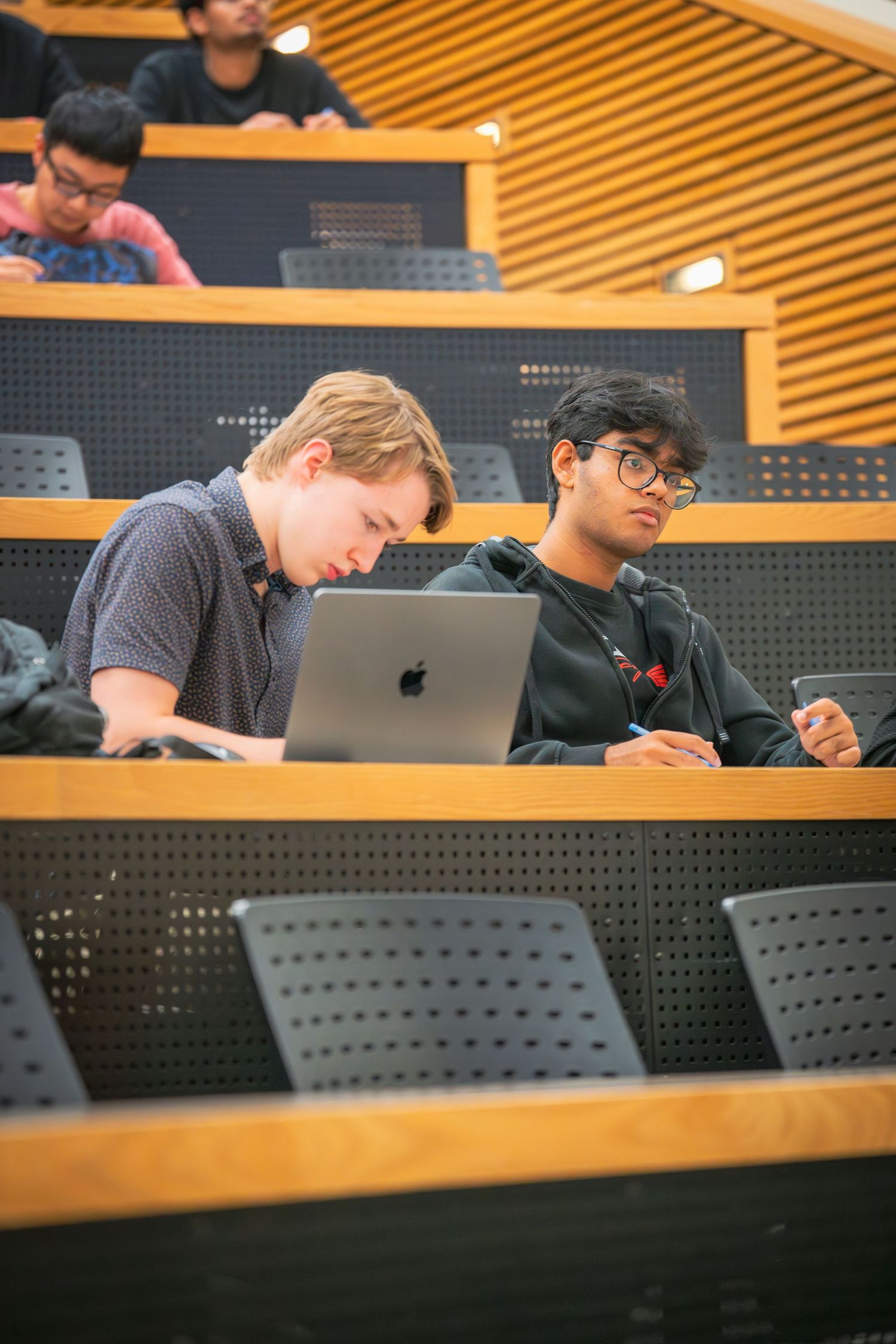 Students working on laptops in tiered seating of a university lecture hall.