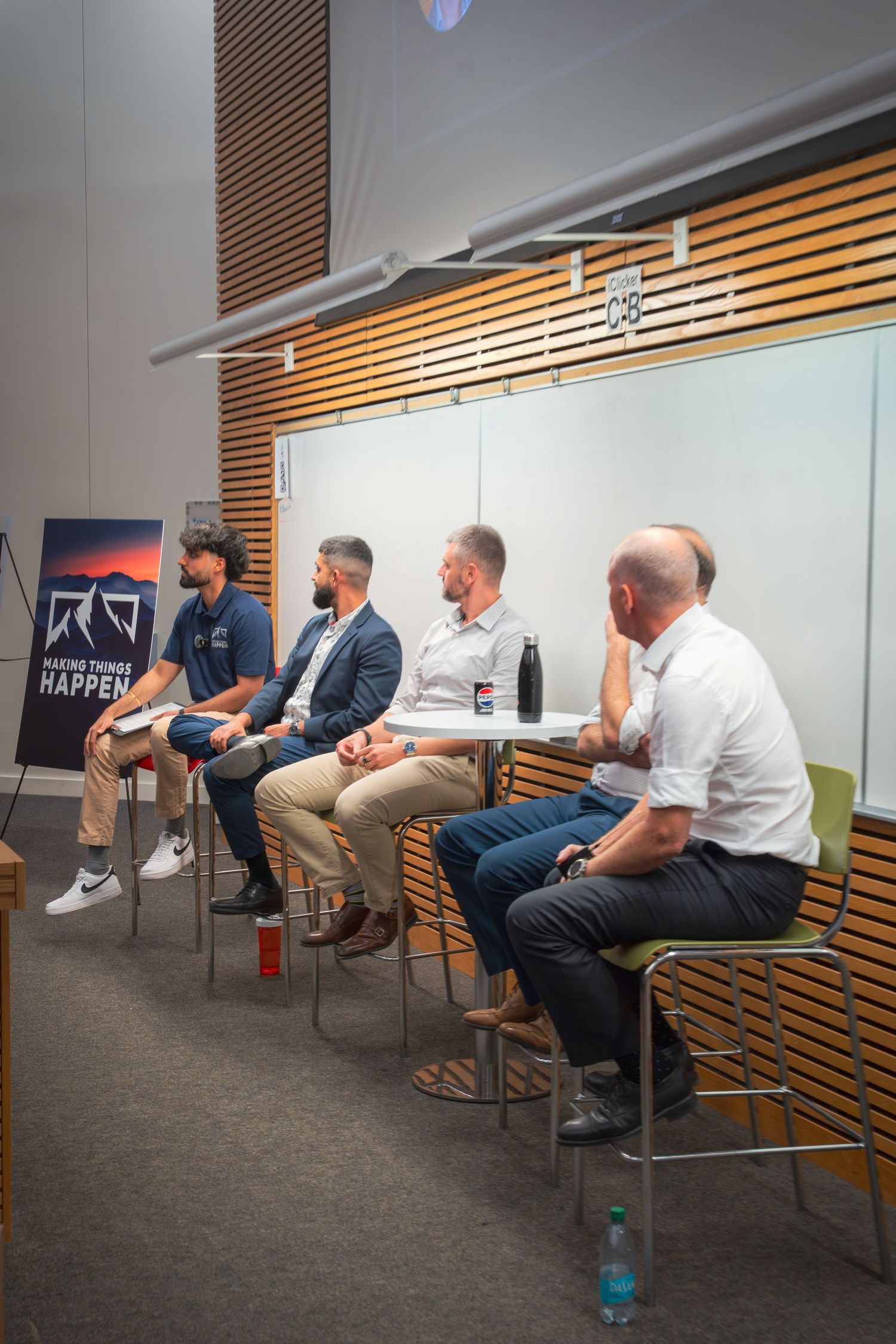 Business professionals seated in chairs during a panel discussion with presentation screen visible.