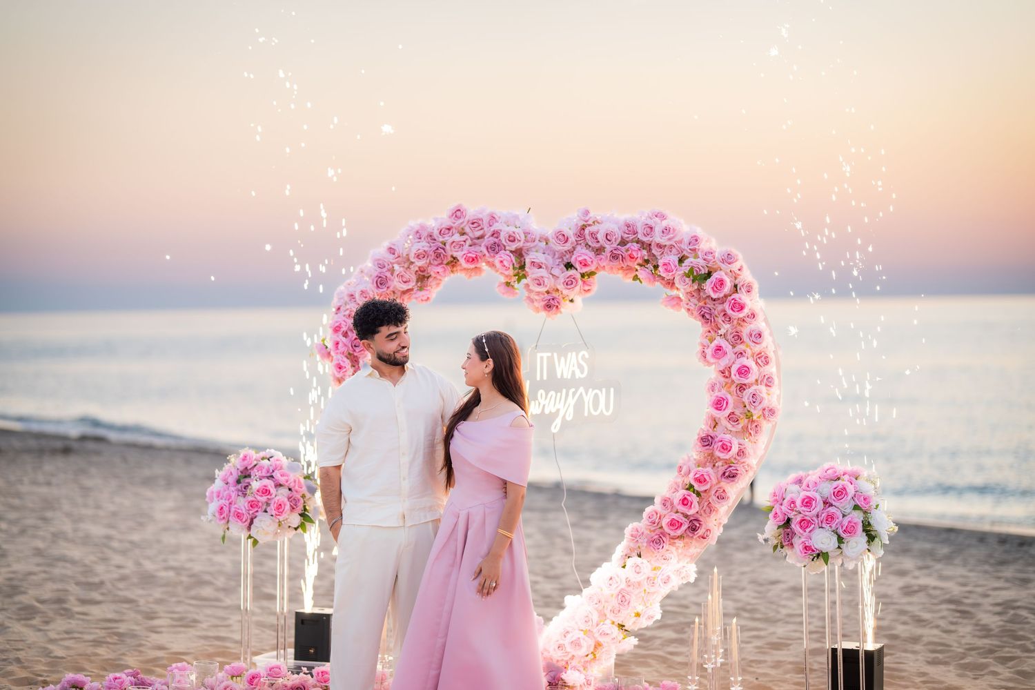 Beach marriage proposal with heart-shaped flower arch and sunset backdrop.