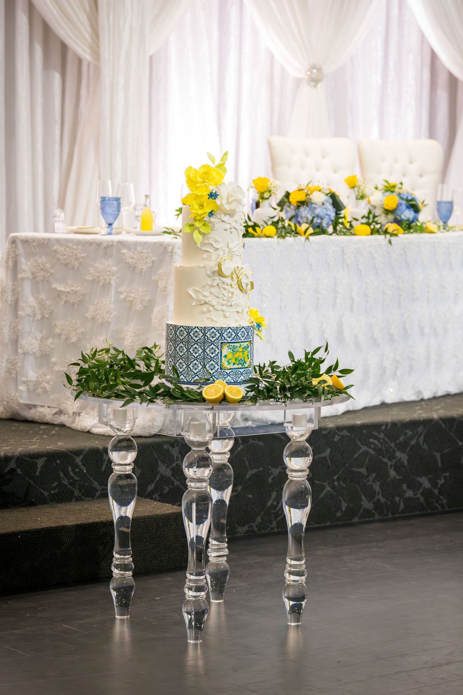 Elegant three-tier wedding cake on crystal stand with blue and yellow floral decor at a wedding reception.