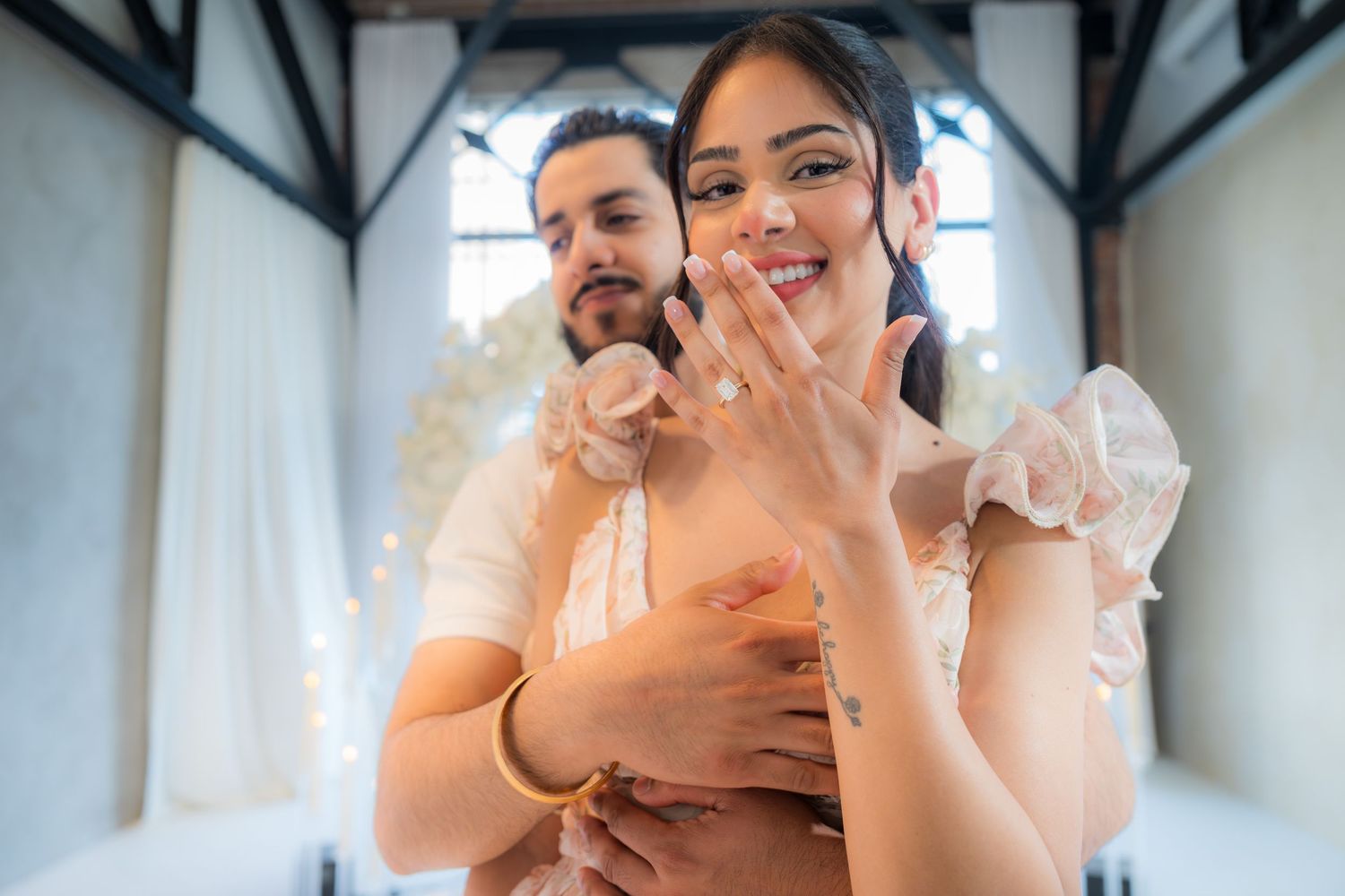 Smiling woman shows off her engagement ring while her partner embraces her from behind.
