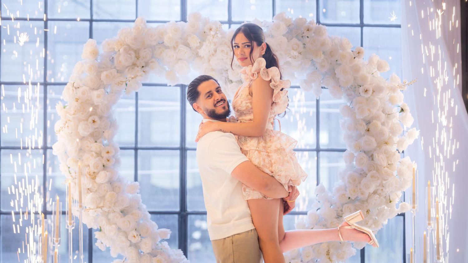 Couple posing happily in front of a white floral heart arch with sparkling fireworks display.