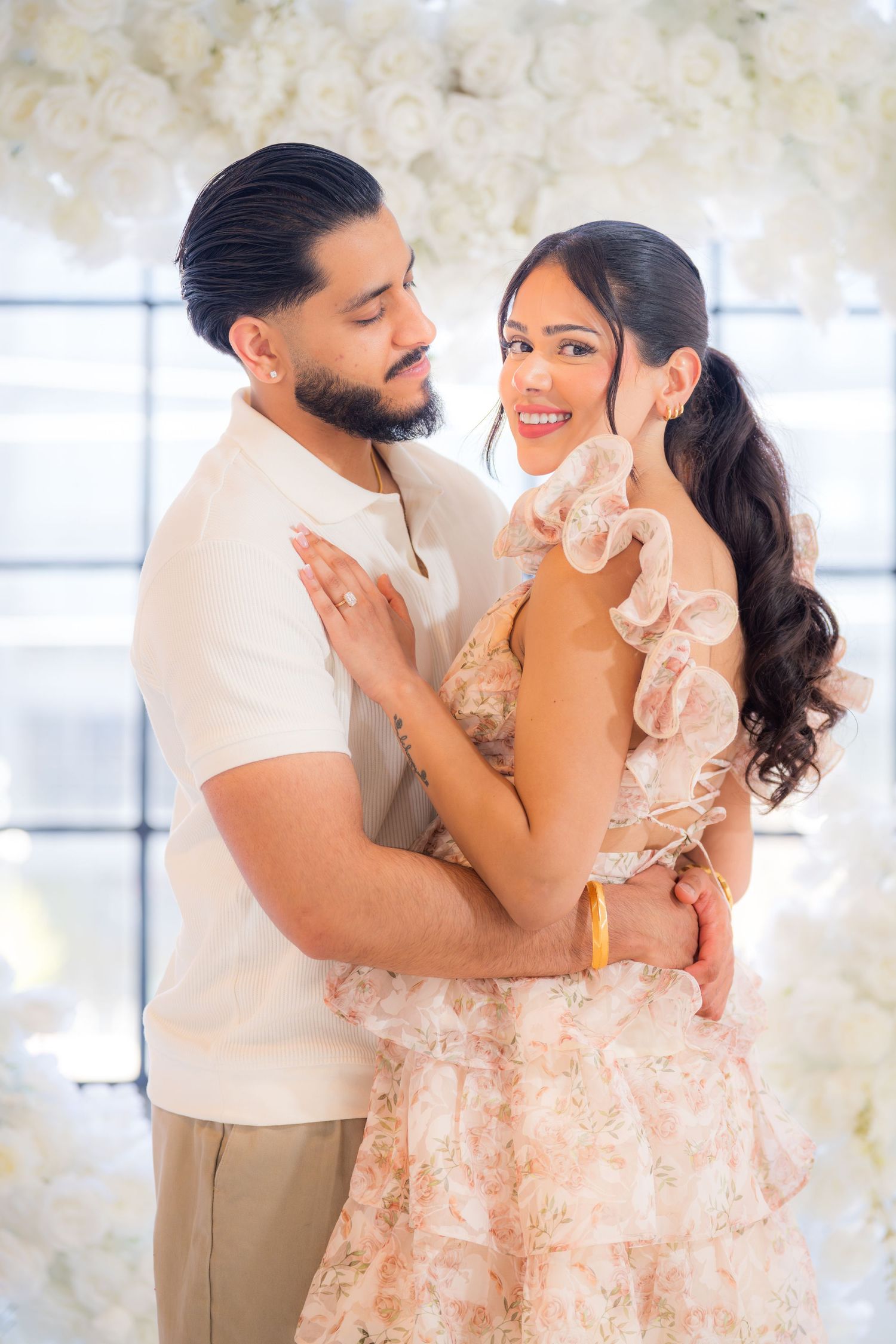 Couple embracing and smiling before a white flower backdrop at engagement shoot.