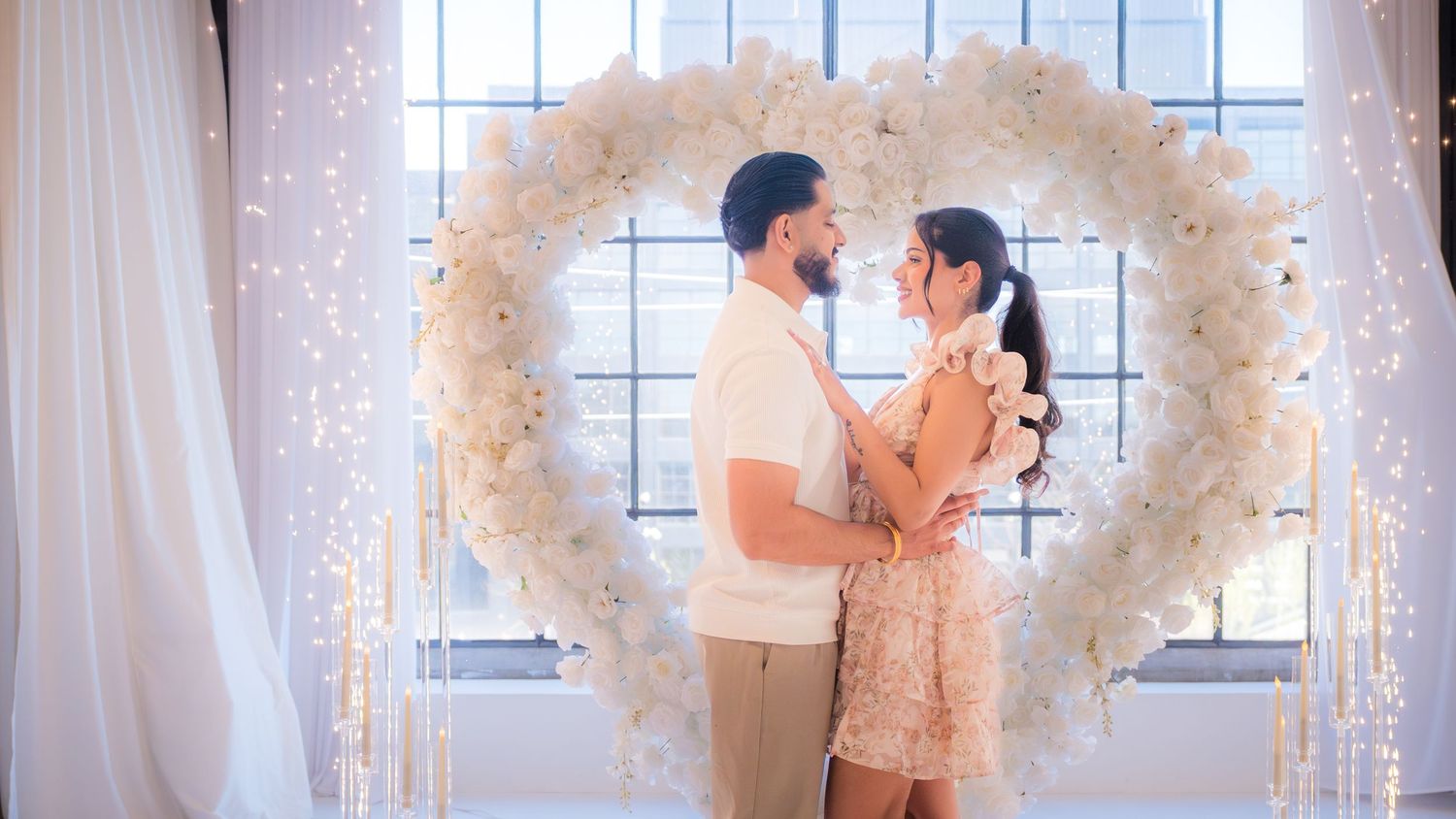 Couple standing close together before white floral heart arch with glowing fairy lights backdrop.