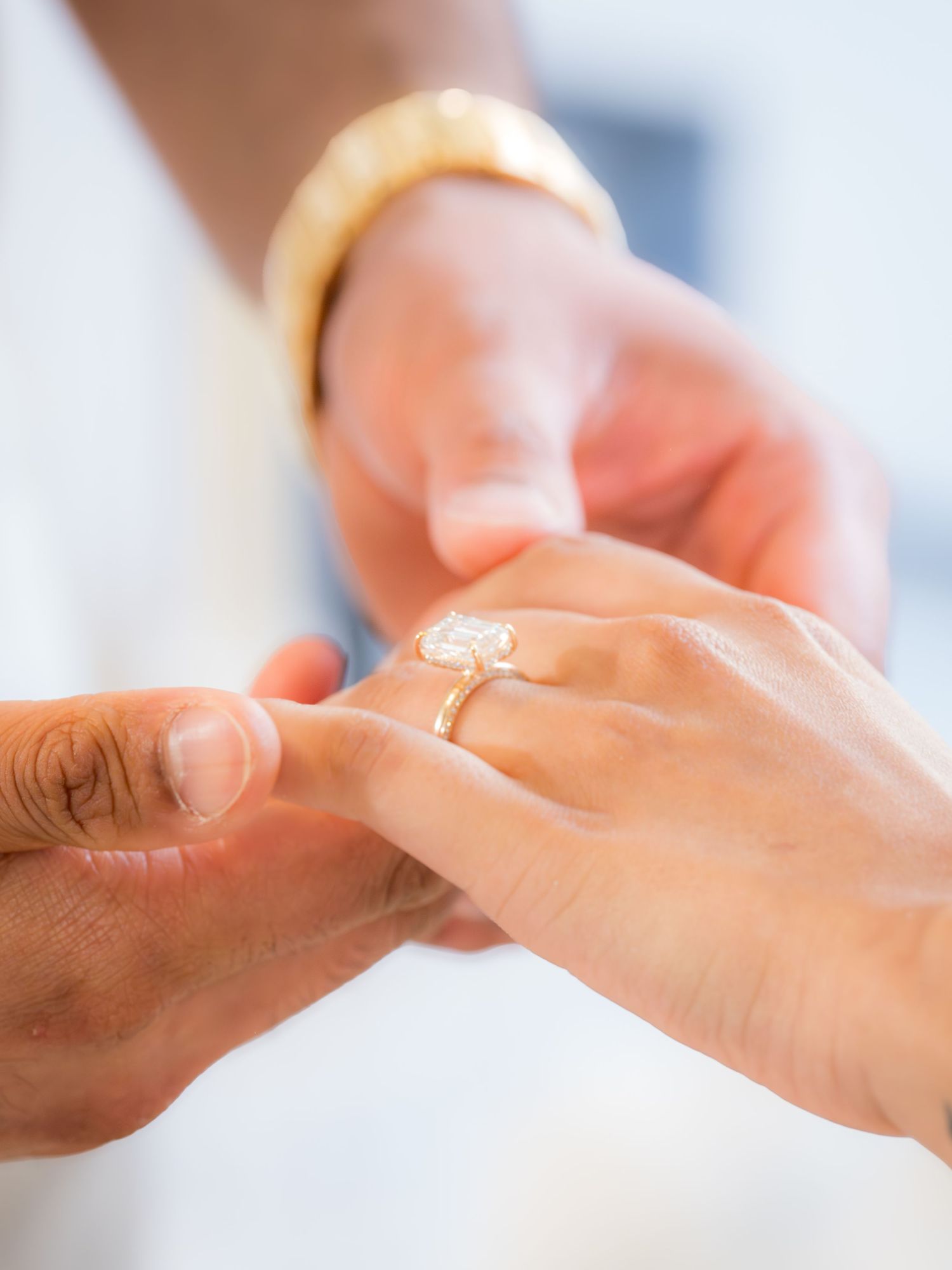 Close-up of diamond engagement ring being placed on bride's finger by groom wearing gold watch.