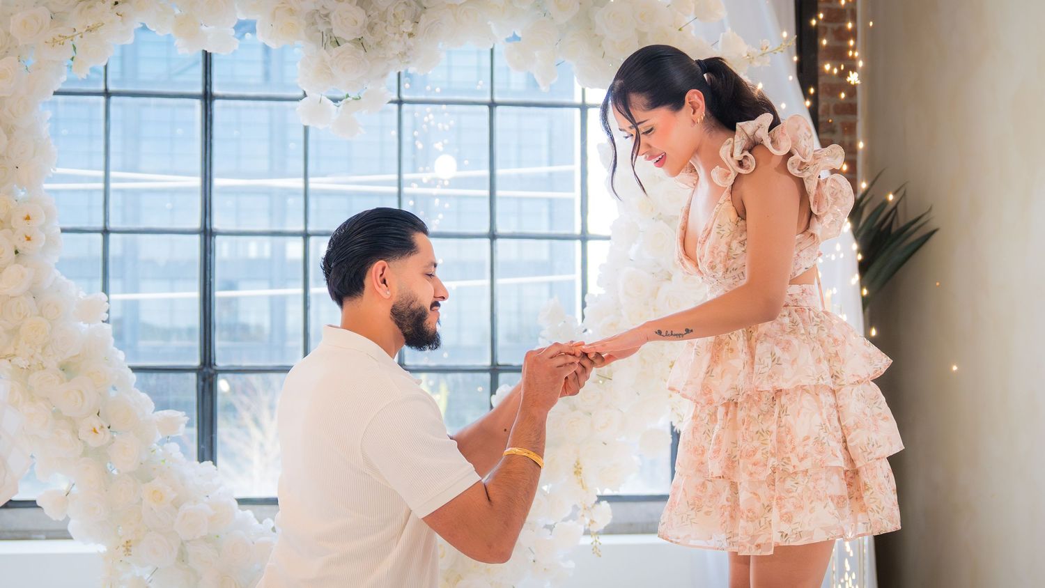 A man kneels on one knee proposing to a woman in a white ruffled dress surrounded by floral decorations.