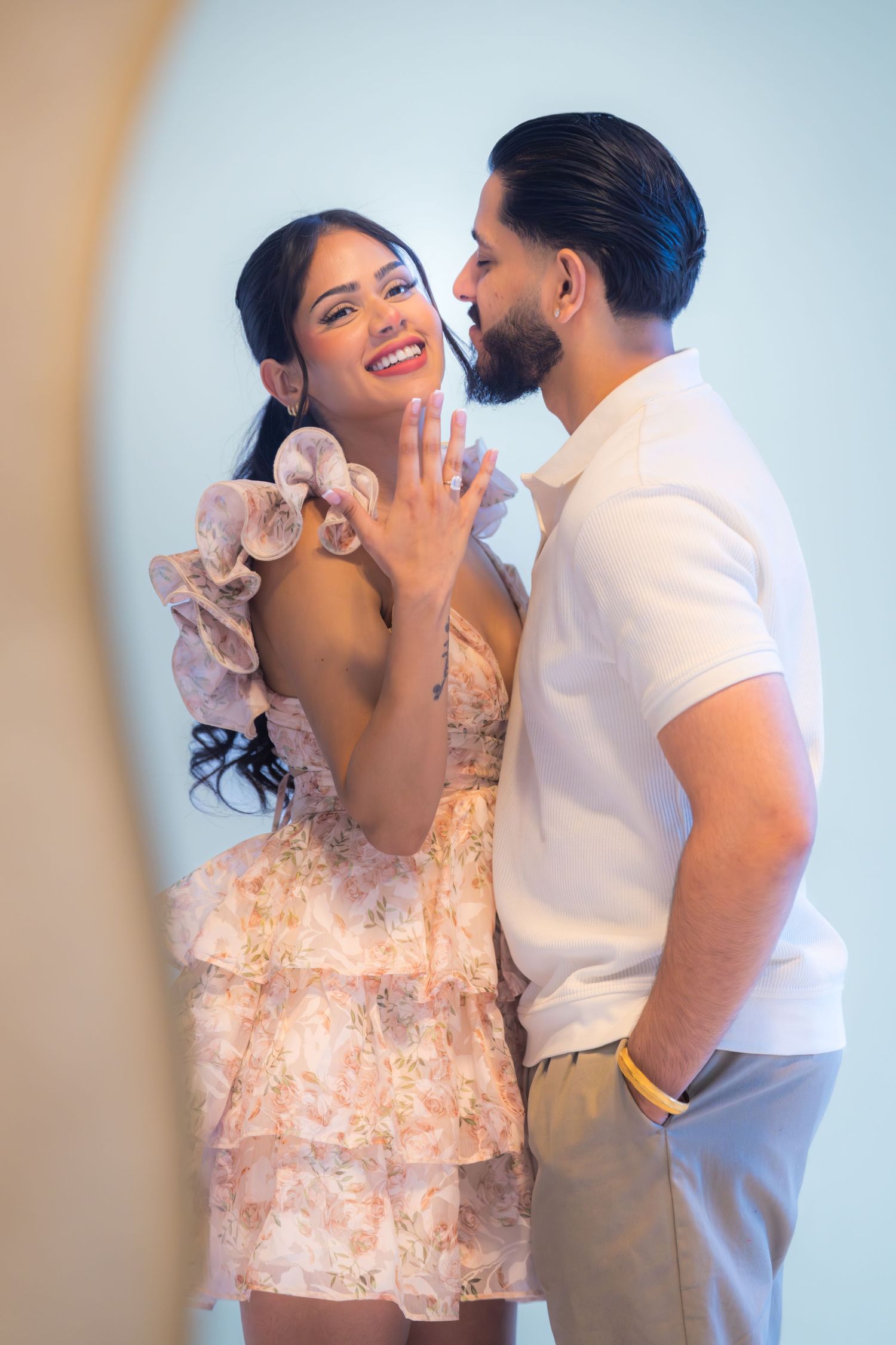 Excited bride-to-be showing engagement ring while partner whispers in her ear at photoshoot.