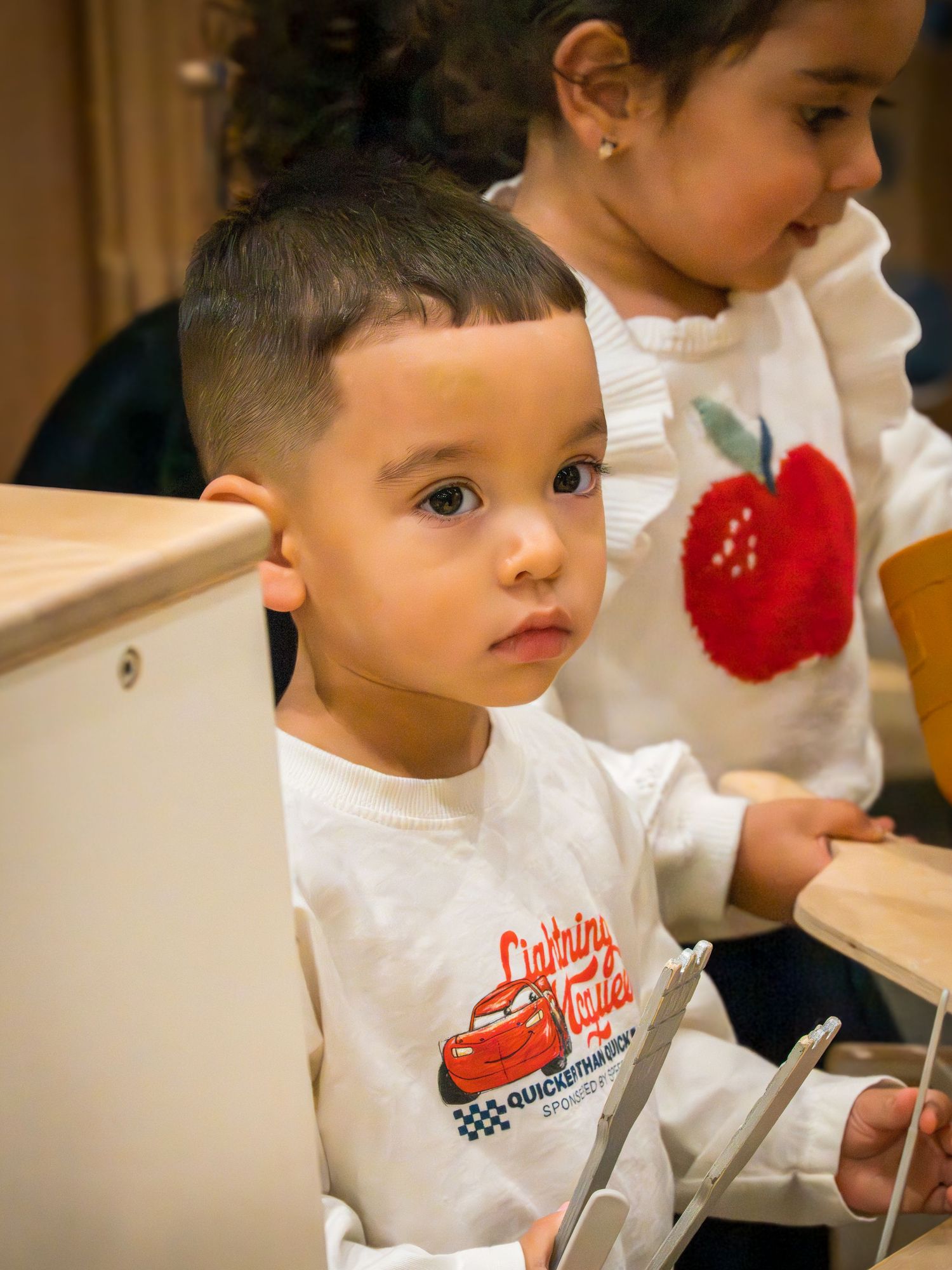 Toddler boy in Lightning McQueen shirt sitting at wooden table with girl in apple sweater.