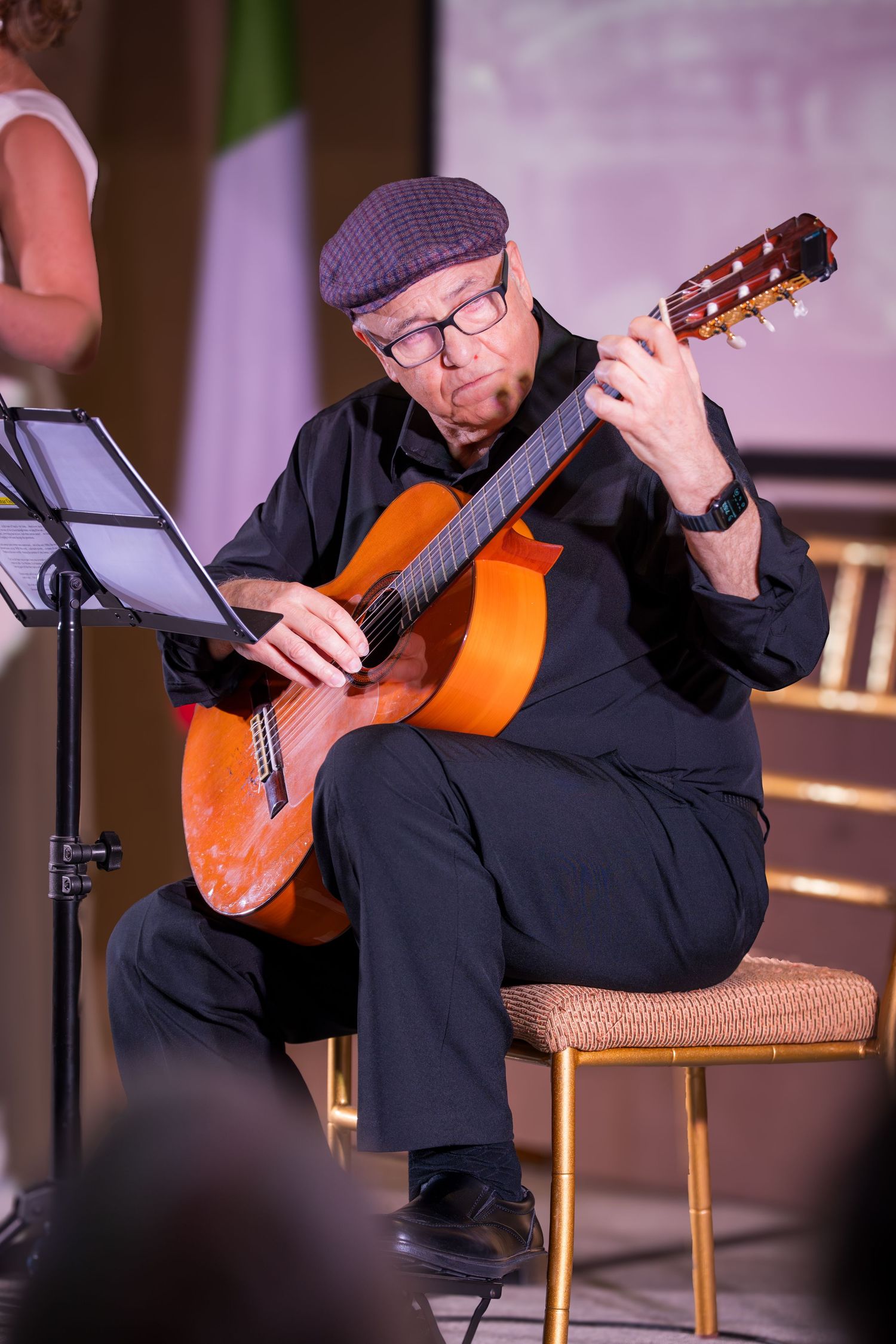An older man in black outfit and cap plays acoustic guitar while reading sheet music on stage.