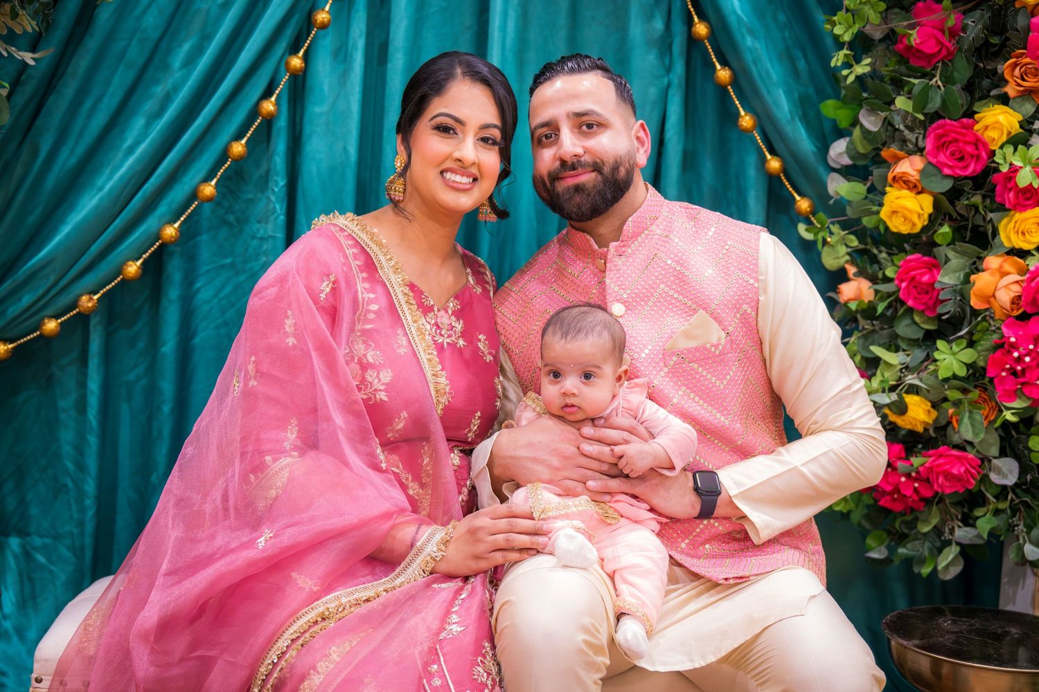 South Asian couple in matching pink outfits holding baby at colorful floral celebration.