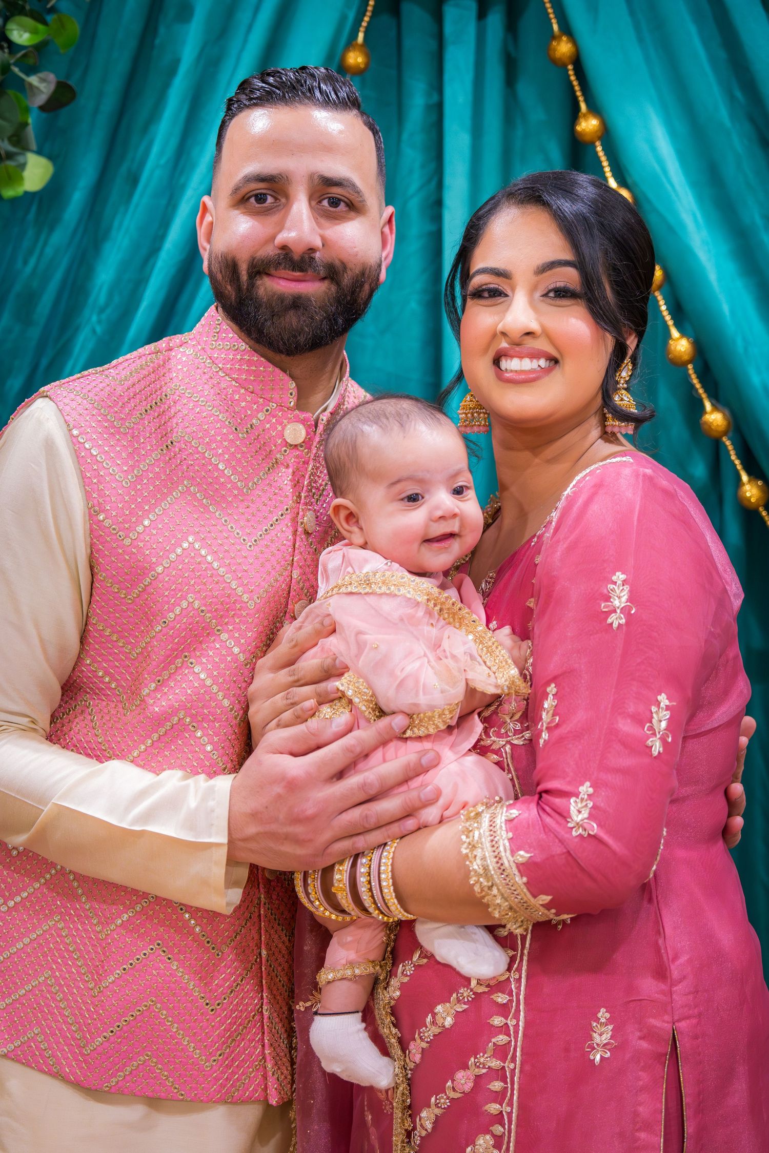South Asian couple in matching pink traditional outfits smiles while holding their newborn baby.