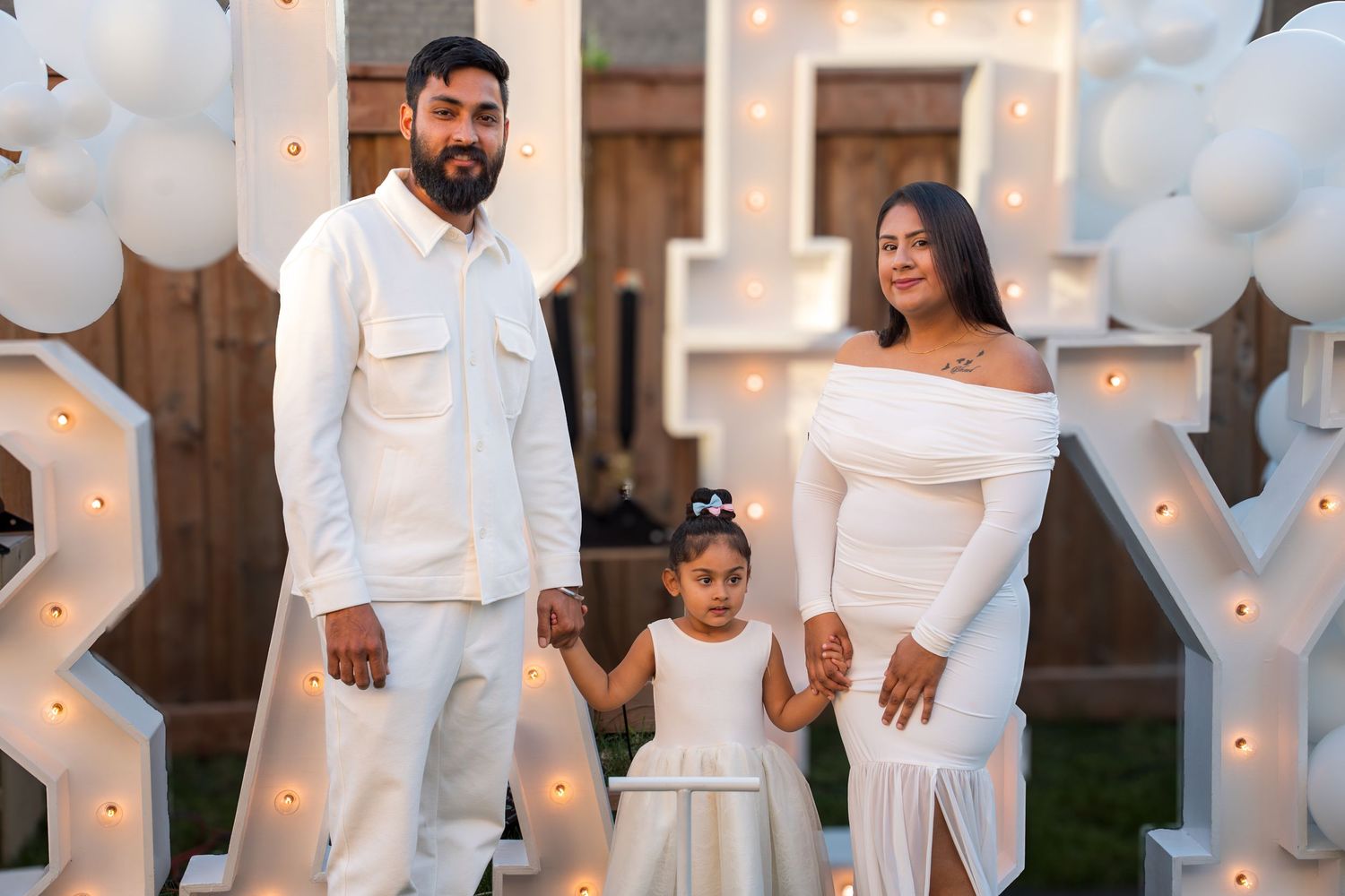 Family of three dressed in white posing together at an elegant outdoor celebration with illuminated letter decorations.