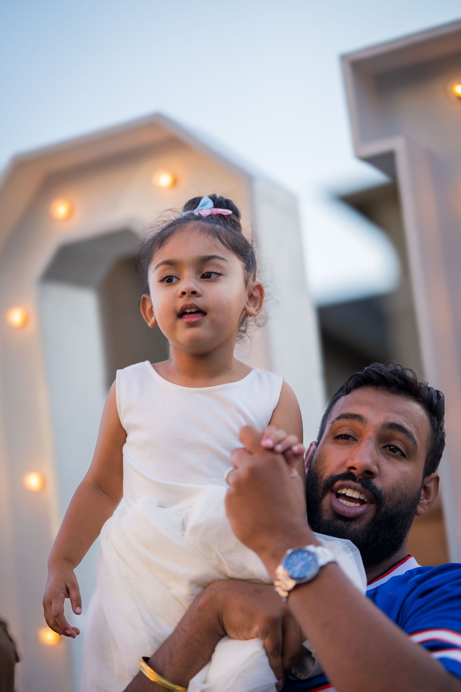 A man holds up a young girl in a white dress at an outdoor celebration with decorative lights.