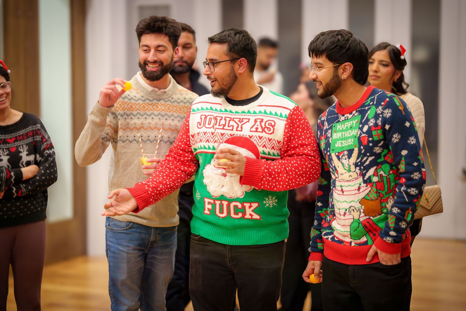 People wearing colorful Christmas sweaters lined up at an indoor holiday party.