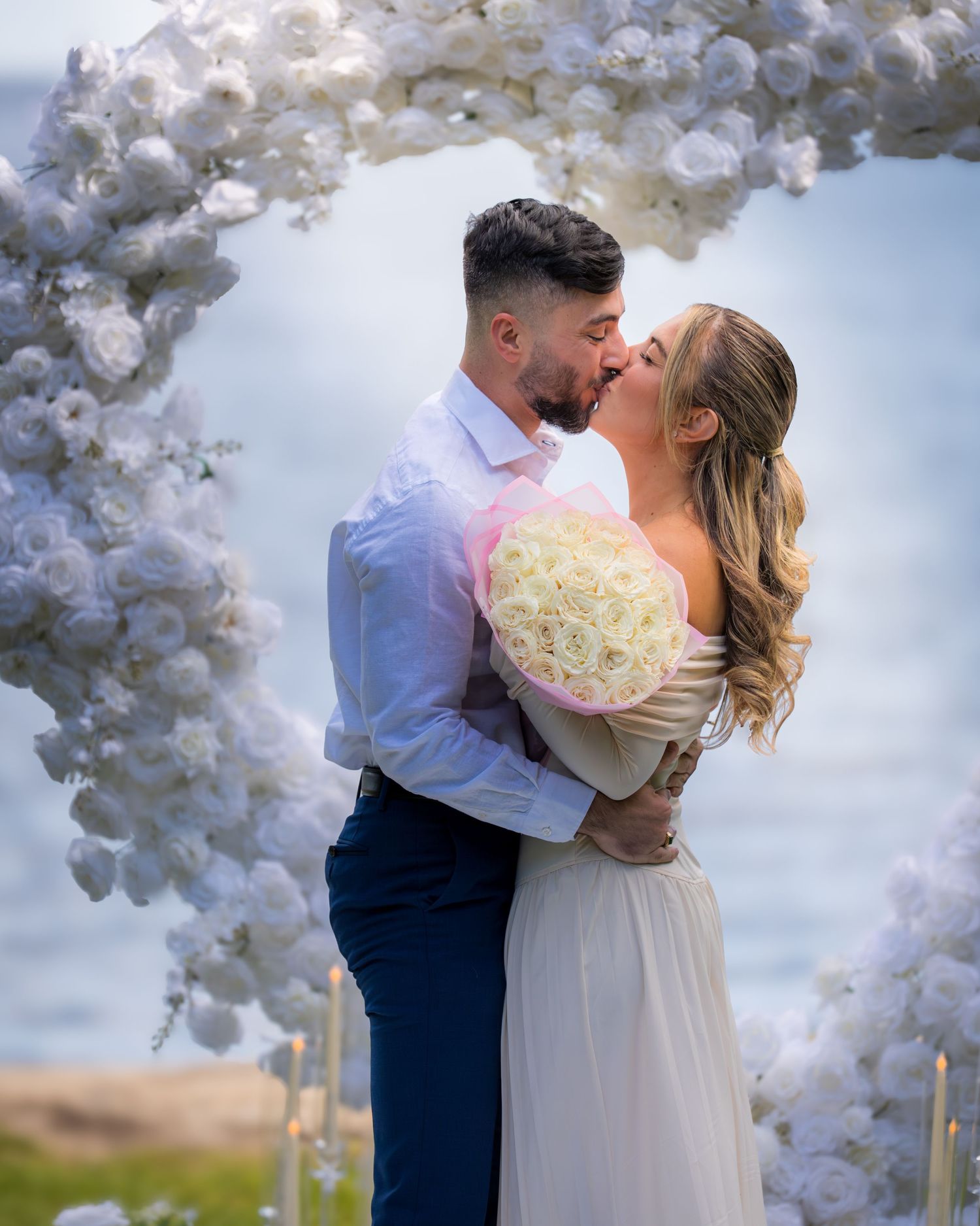 Wedding ceremony under white floral arch with ocean backdrop.