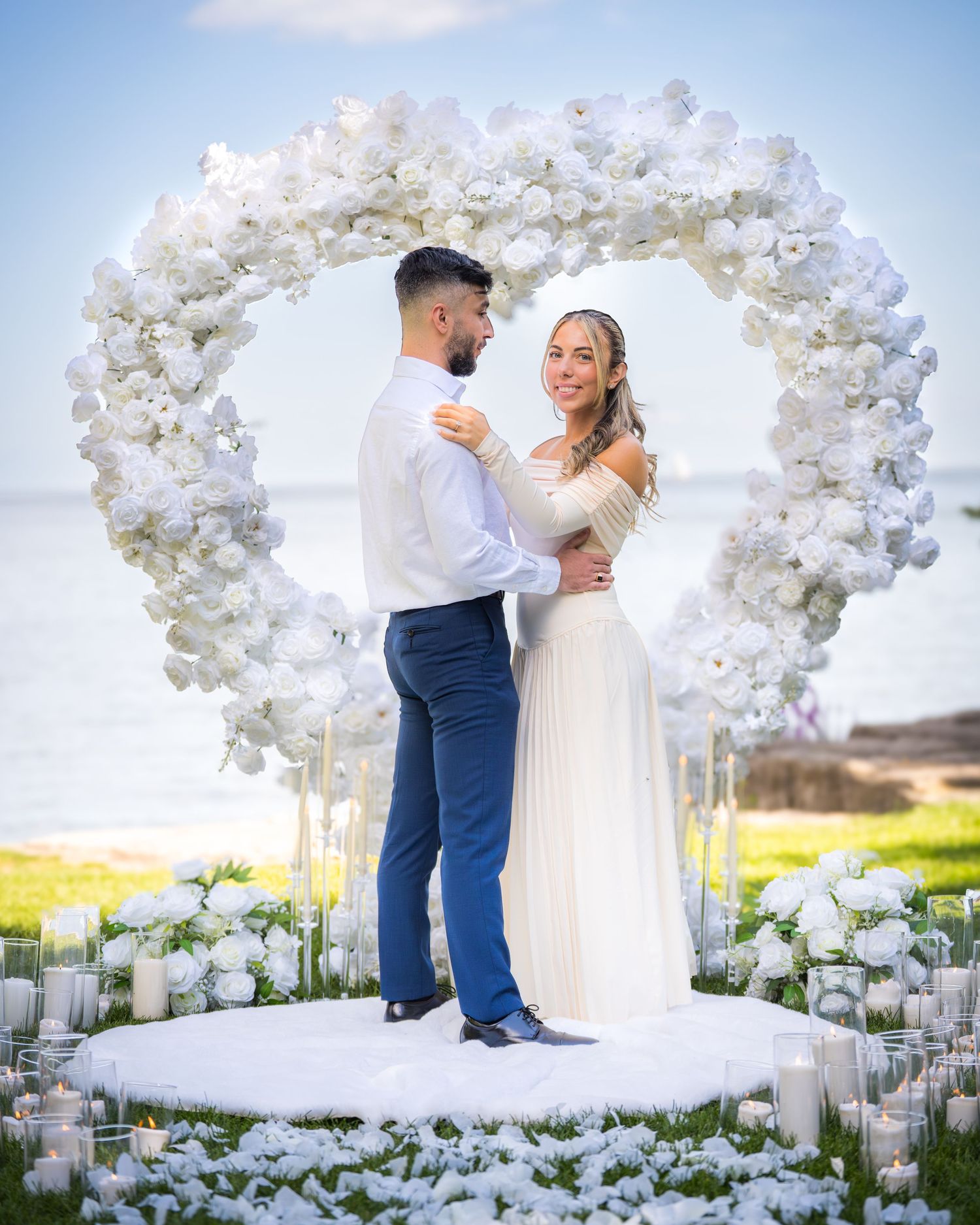 A romantic wedding ceremony setup with white floral arch and candles on a scenic oceanfront lawn.