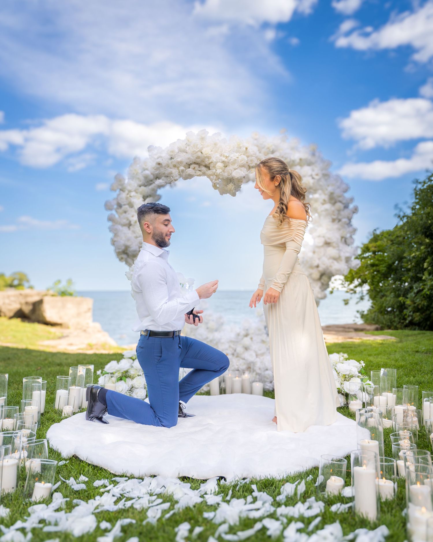 Romantic proposal setup with white floral arch and candles on a grassy lawn under blue skies.