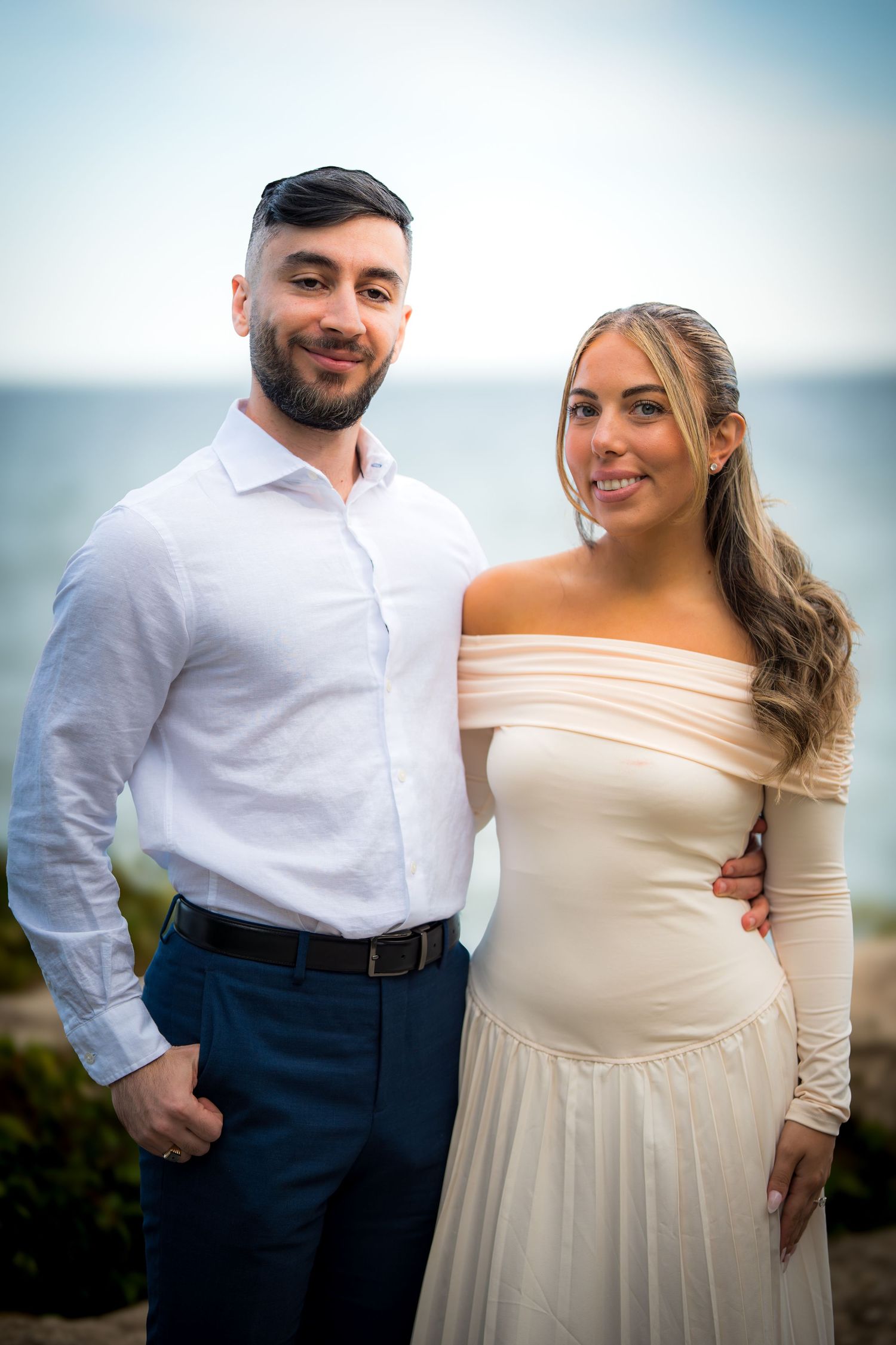 Couple in cream and navy formal wear posing together for seaside portraits.