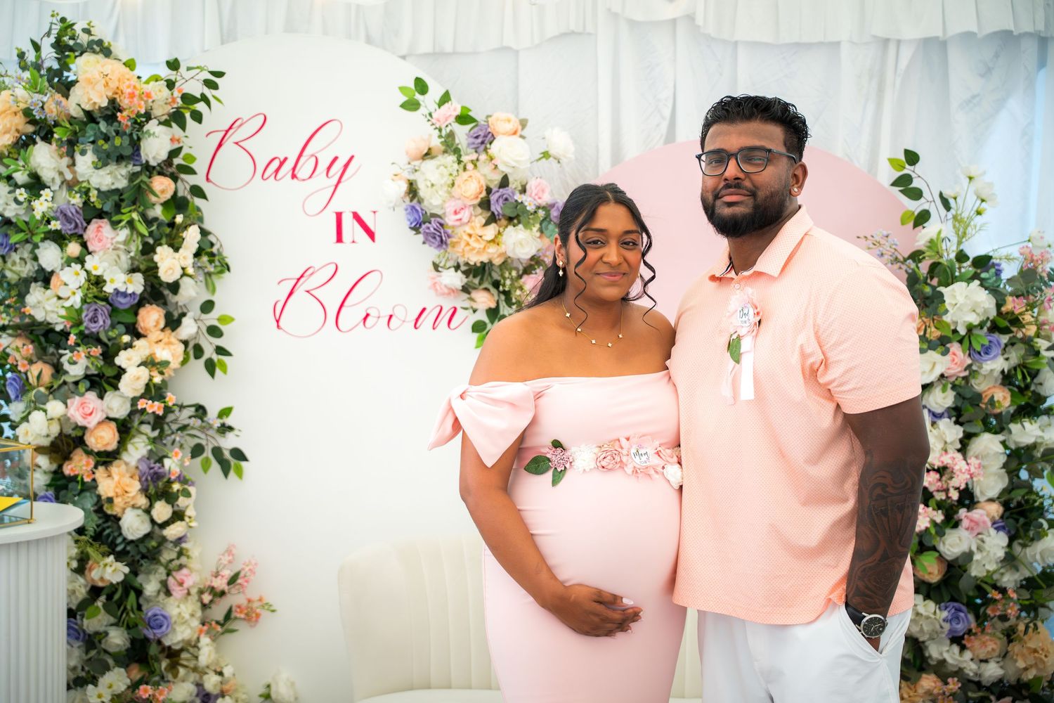Expectant parents sharing a tender moment at their baby shower with floral arch backdrop.