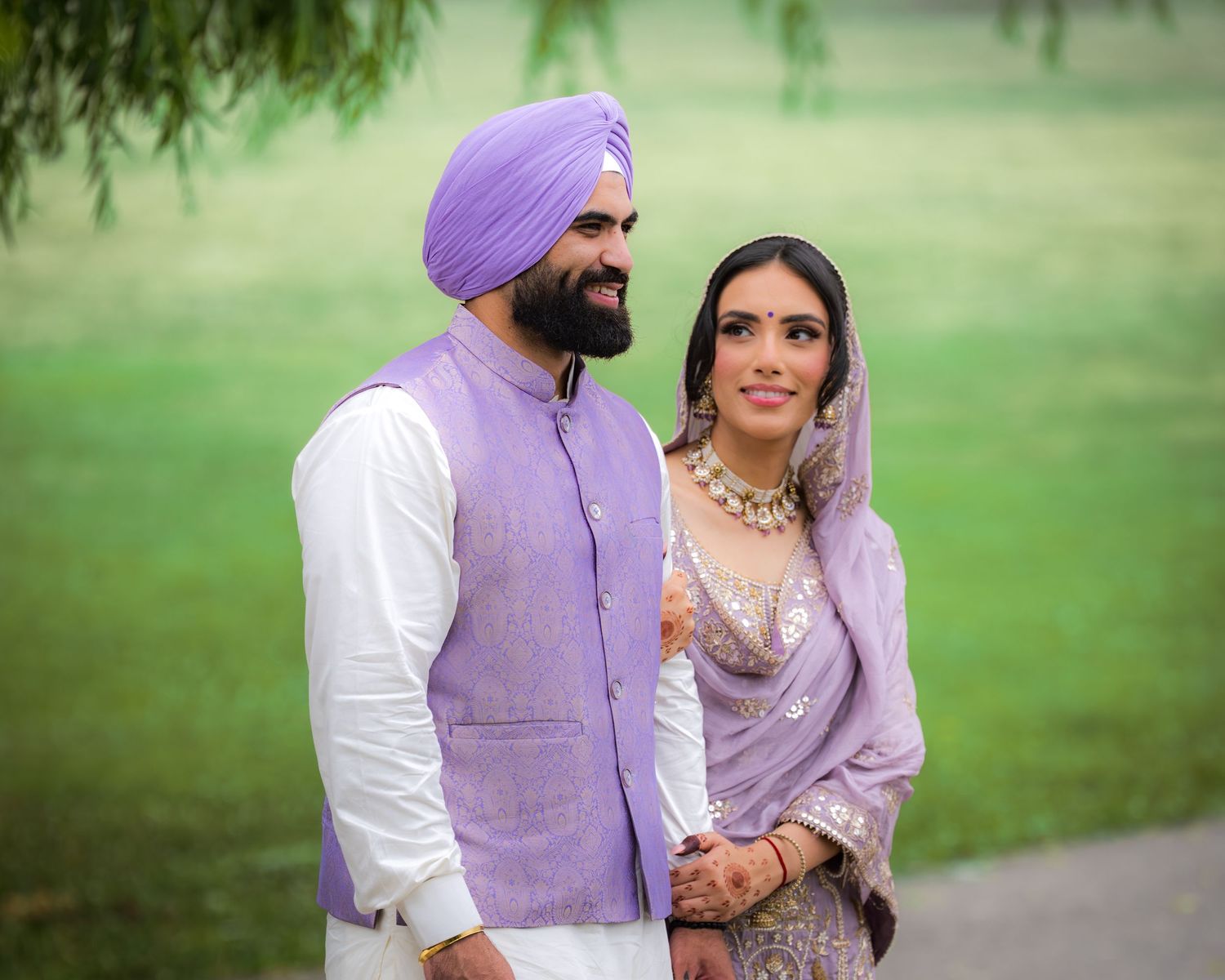 A couple in coordinated purple and white traditional Punjabi attire posing against a lush green background.
